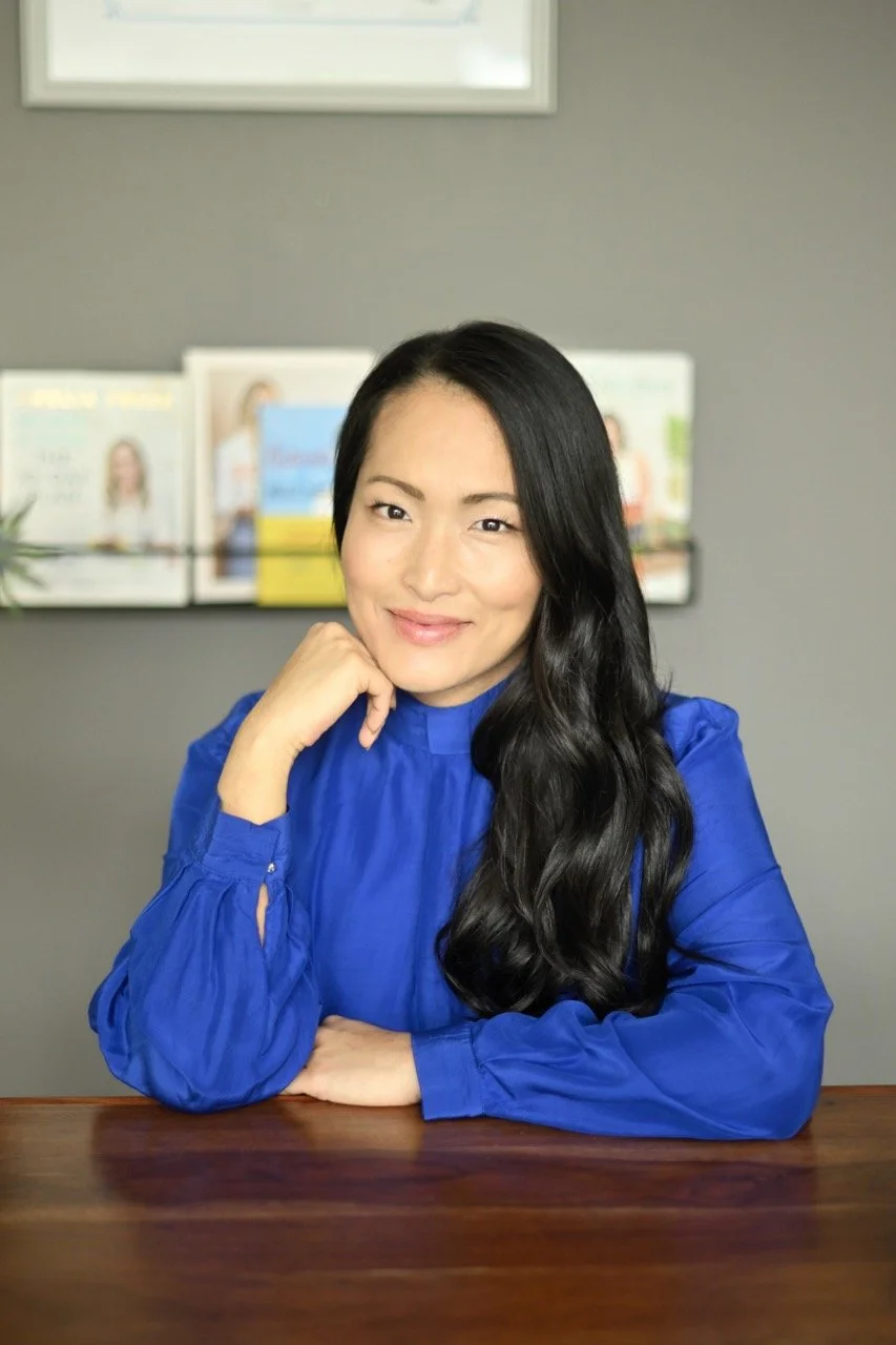 A woman with long black hair wearing a blue blouse sitting at a wooden table, smiling, with magazines or books on a shelf behind her.
