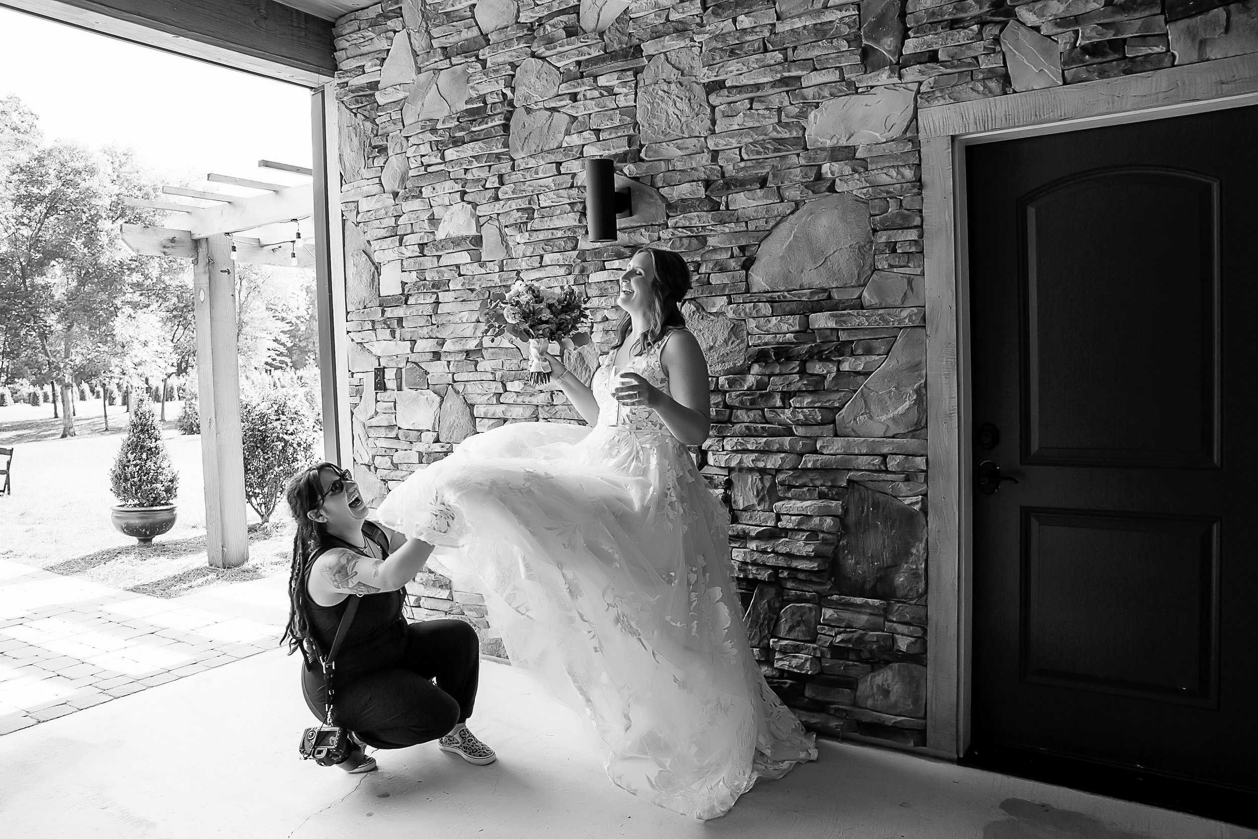 black and white image of photographer laughing while fluffing brides wedding dress
