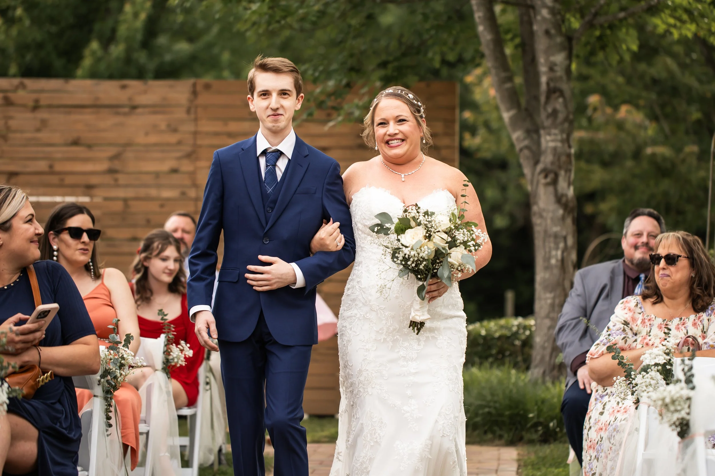 smiling bride being walked down the aisle by her son who is wearing a navy blue suit outdoor summer wedding charlotte nc