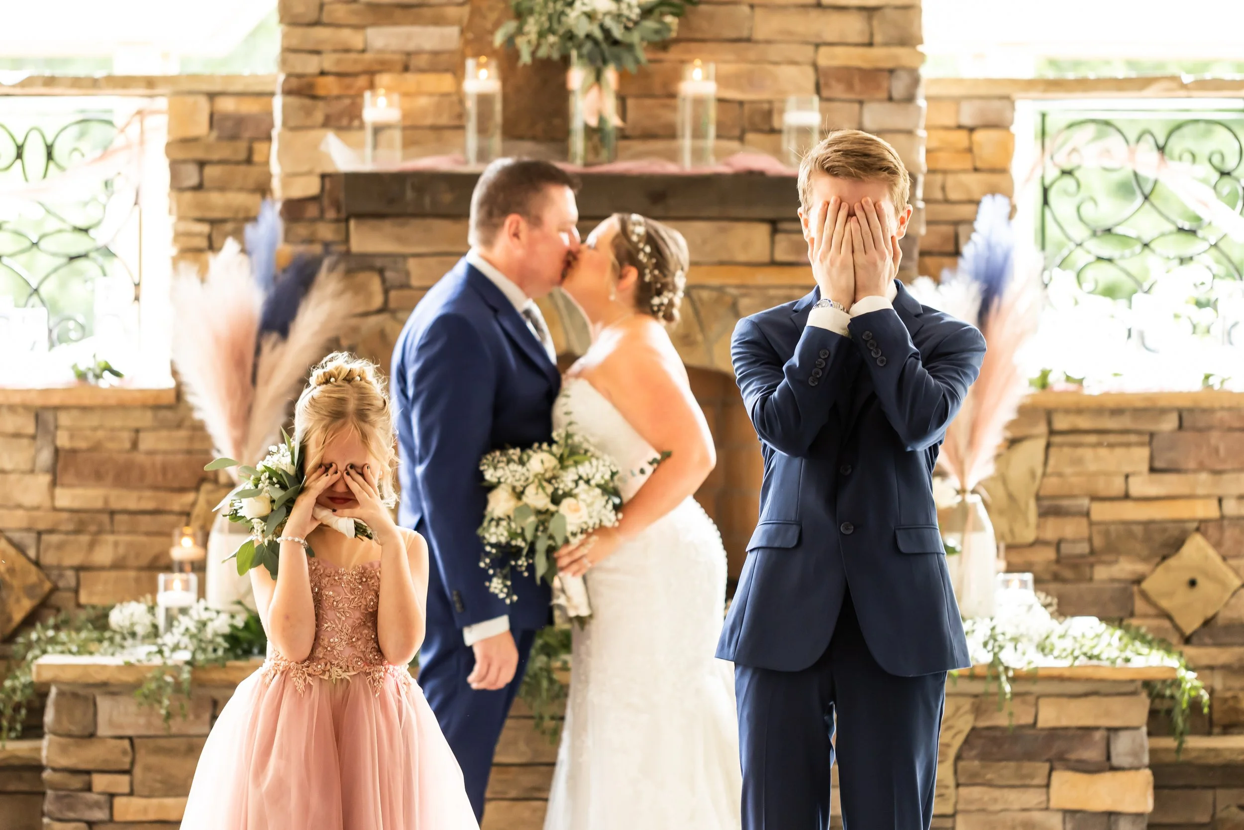 kids covering their faces while mom and dad kiss in the background at their wedding
