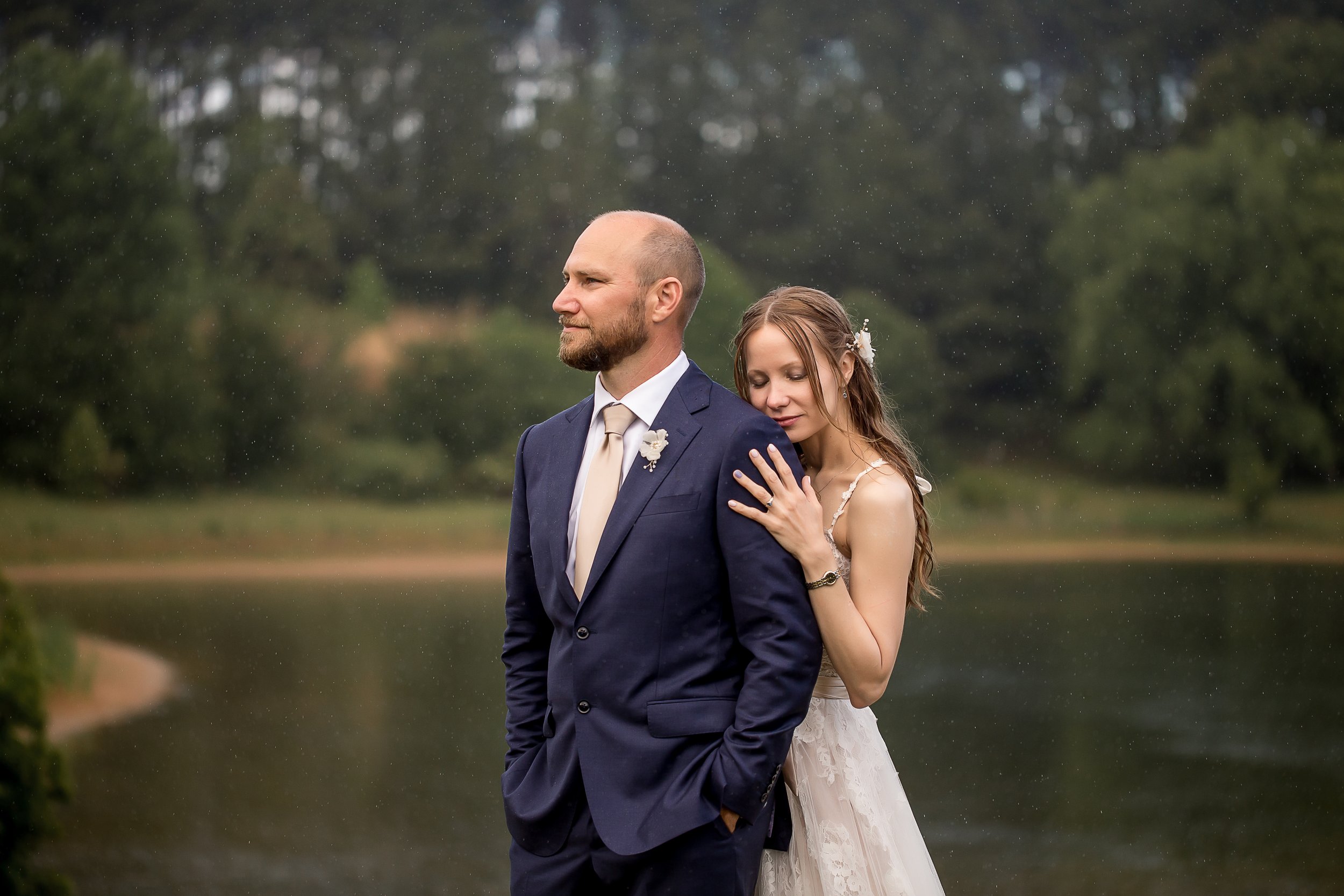 Bride with long red hair and groom in navy suit with blush colored tie stand quietly in the soft rain in front of a lake.