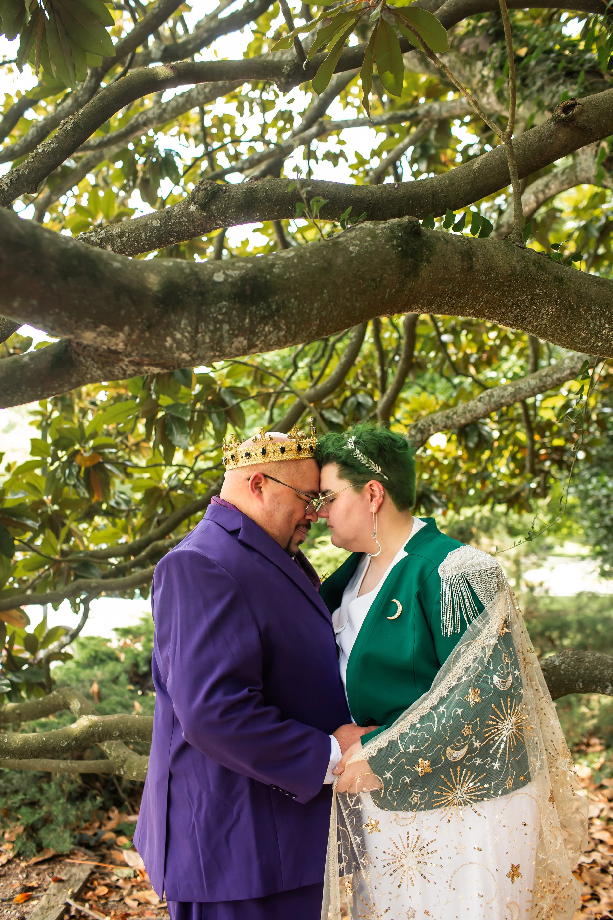 unique couple with royal purple suit and crown and green hair with gold cape embrace under large magnolia tree after eloping