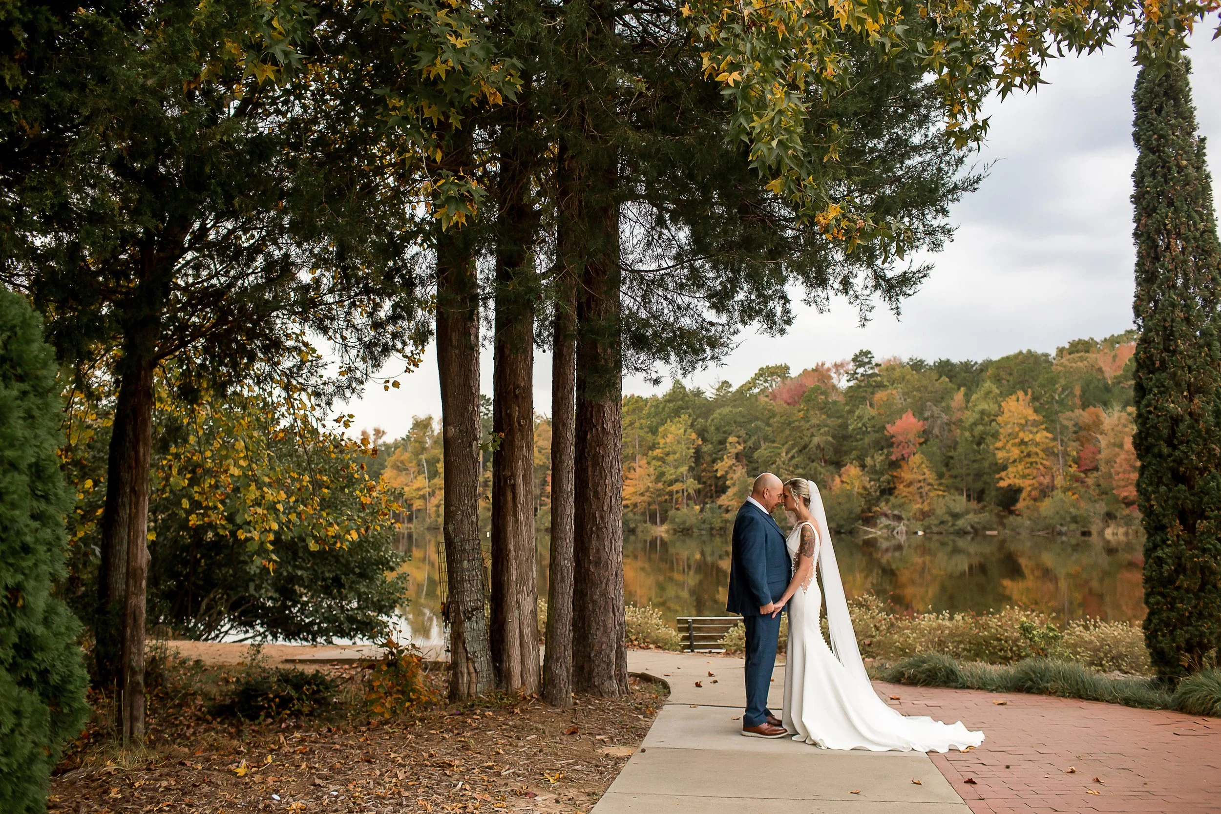 bride and groom embrace udnerneath tall trees with fall foliage