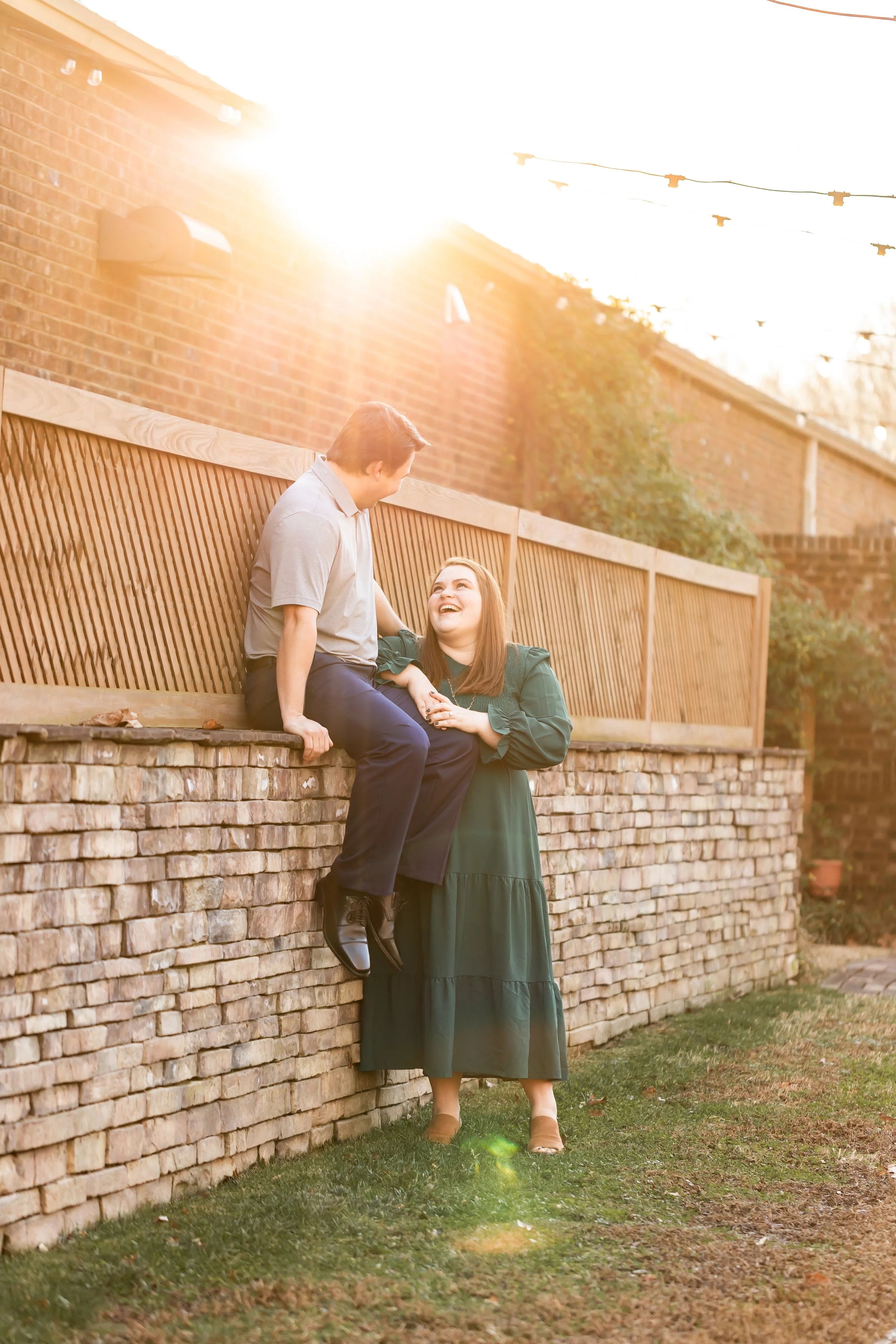 laughing woman in green dress with fiance sitting on brick wall golden light pouring in charlotte engagement photographer