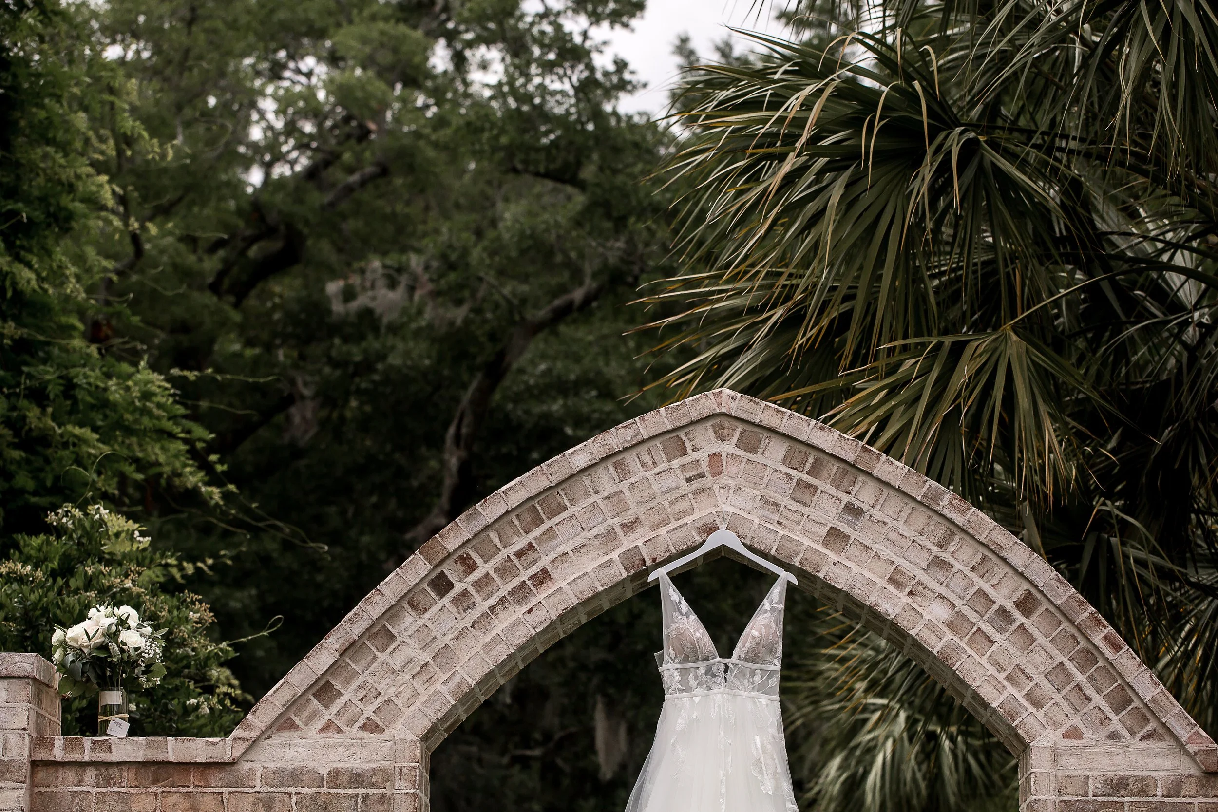 wedding detail photo of dress and bouquet in brick arch with greenery around it