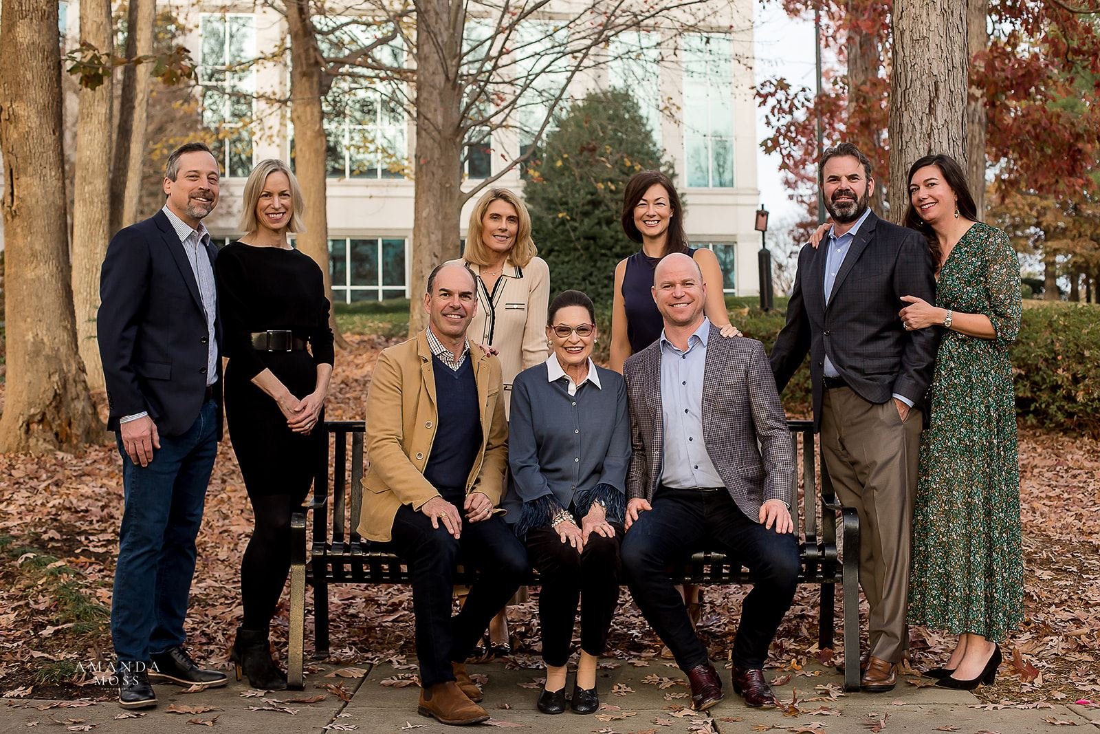 family photo in the fall with mom and adult sons sitting on park bench and four more adults standing around them smiling