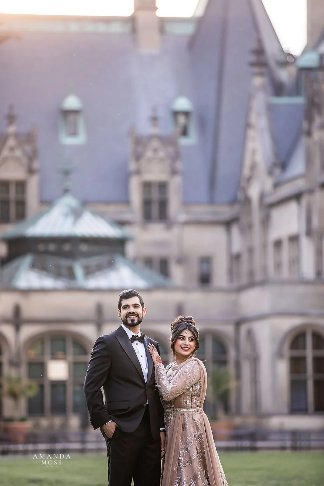 Indian wedding couple embraces and looks off into distance in front of biltmore estate. they are wearing a tuxedo and gold dress