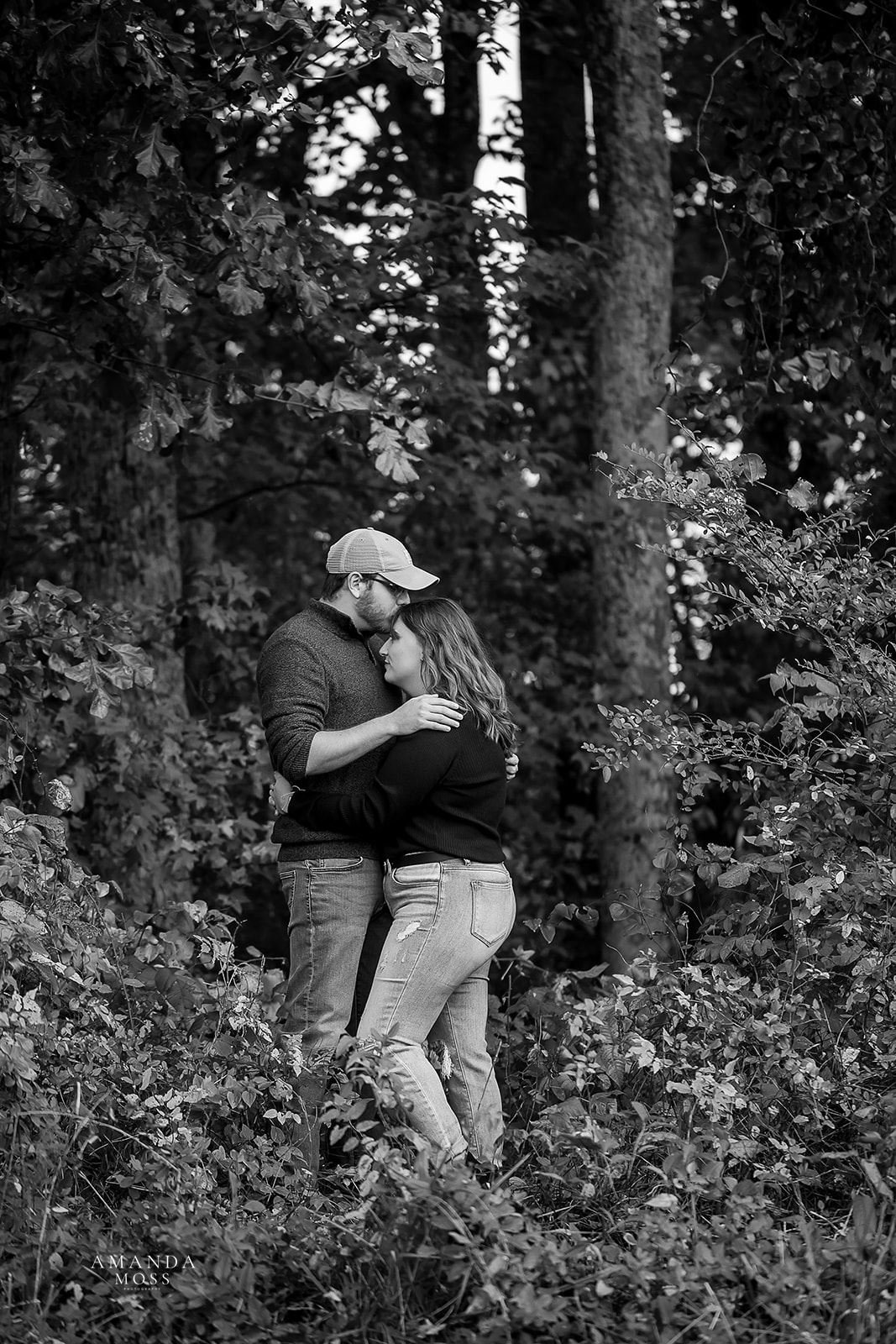 black and white photo of couple embracing while the man kisses the woman on the forehead they are standing in the middle of the woods