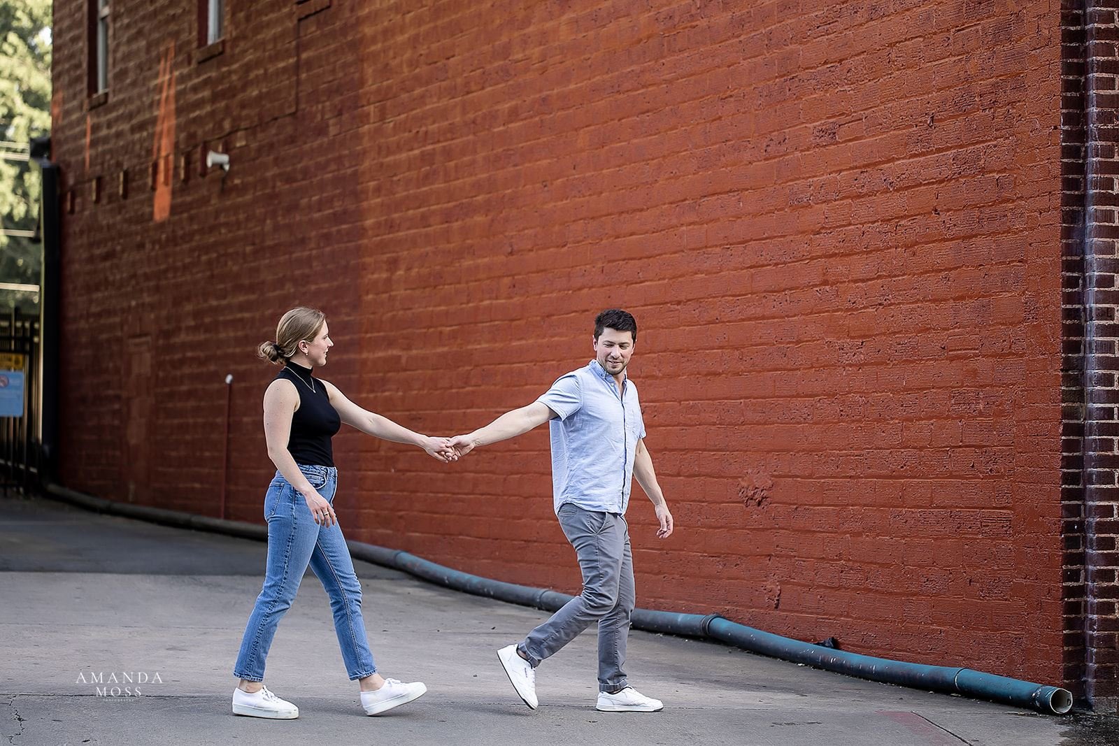 man leads fiance on walk across the street in front of a large red brick wall during outdoor engagement session in noda charlotte nc