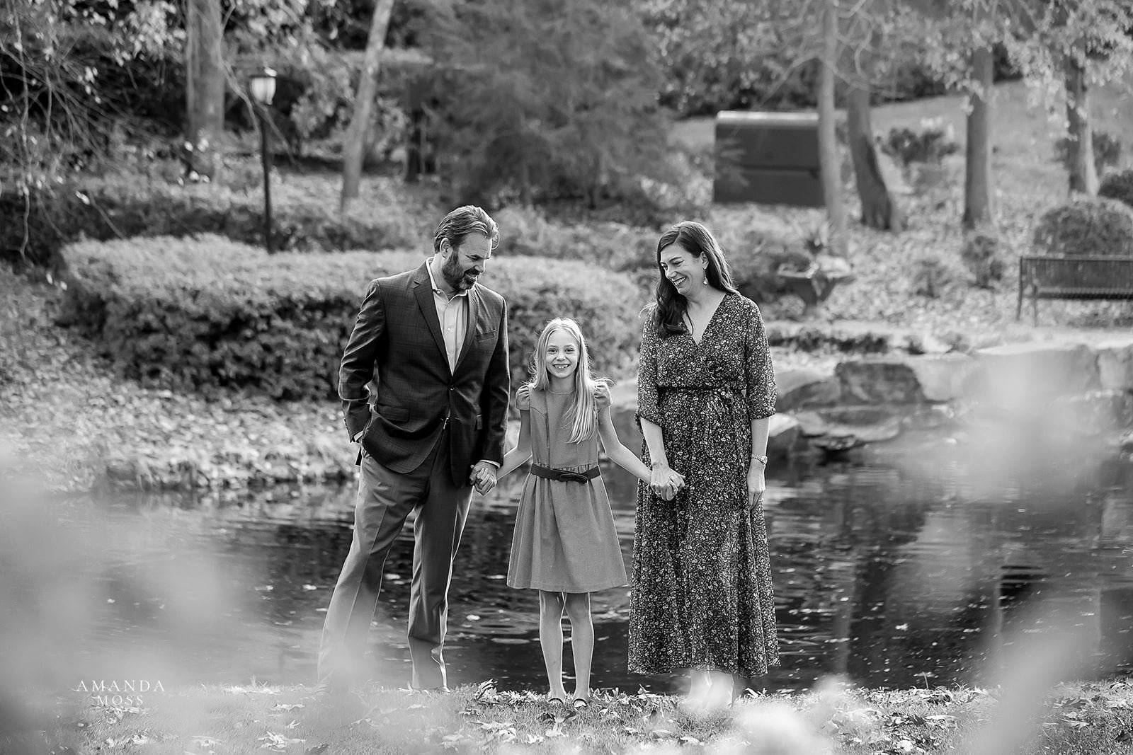 black and white family photo of parents holding their ten year old daughters hand while walking towards the camera with a pond in the background