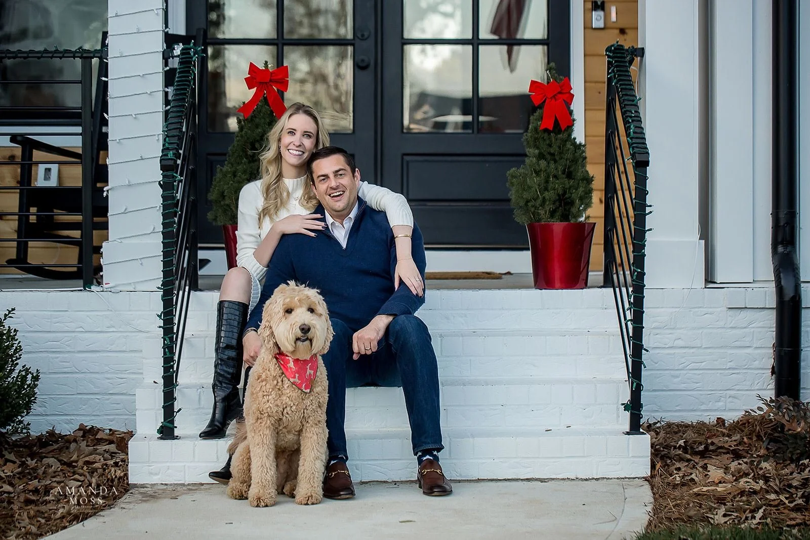 smiling couple with arms around each other sit on front porch of new home with their labradoodle in a red scarf