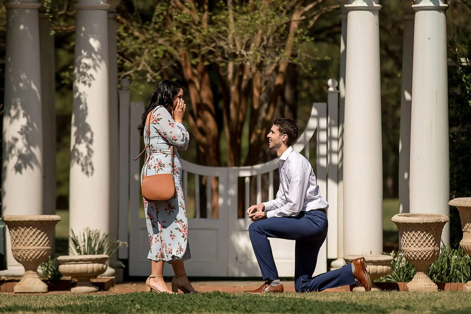 man in navy pants proposes to girlfriend in light blue floral dress at botanical garden