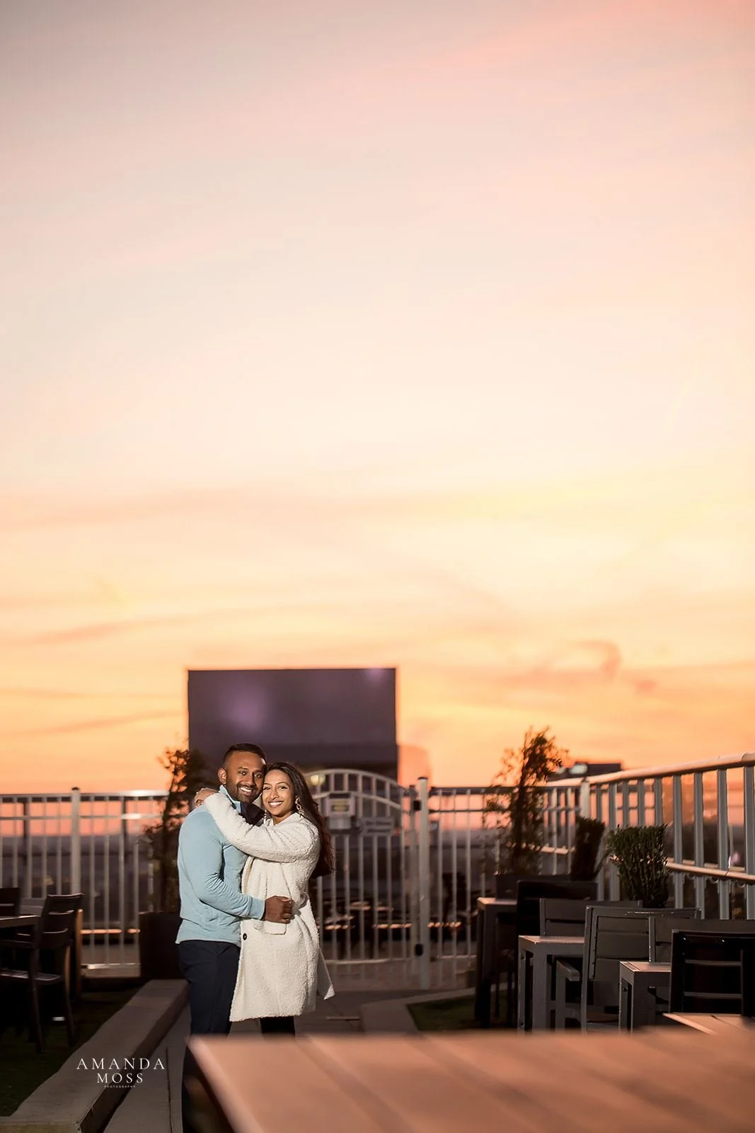 newly engaged couple poses with arms around each other with a fiery sunset in the background