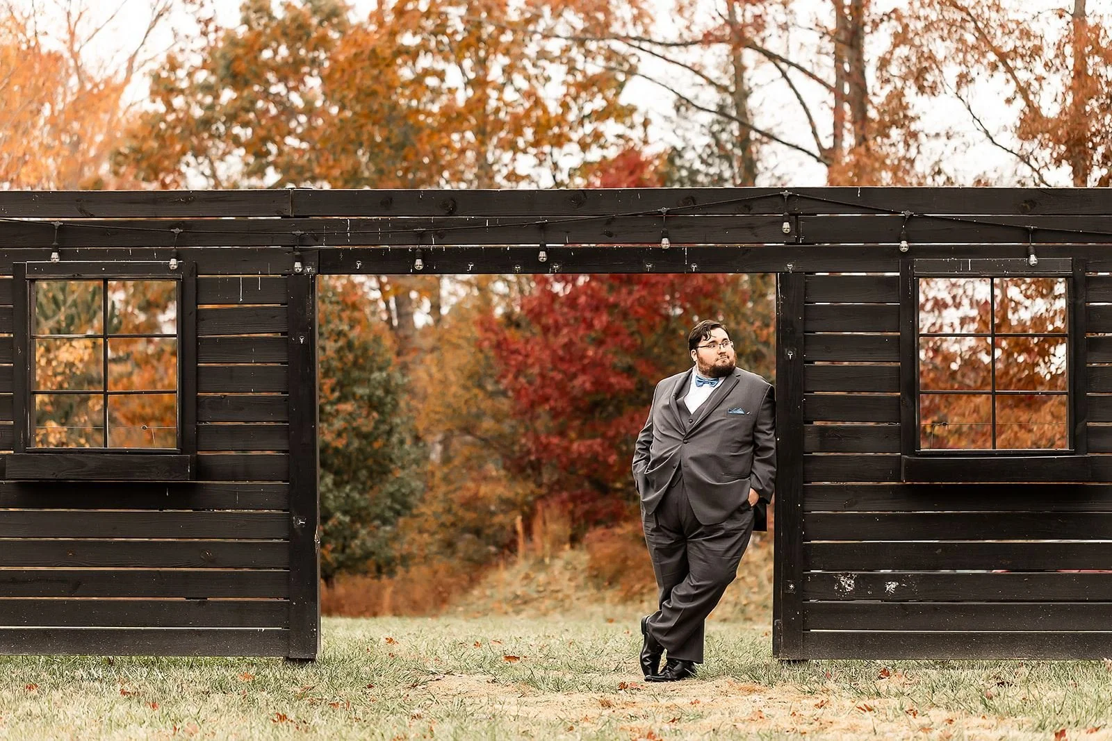 groom leans against wooden frame at bansen farms wedding venue in north carolina fall foliage behind