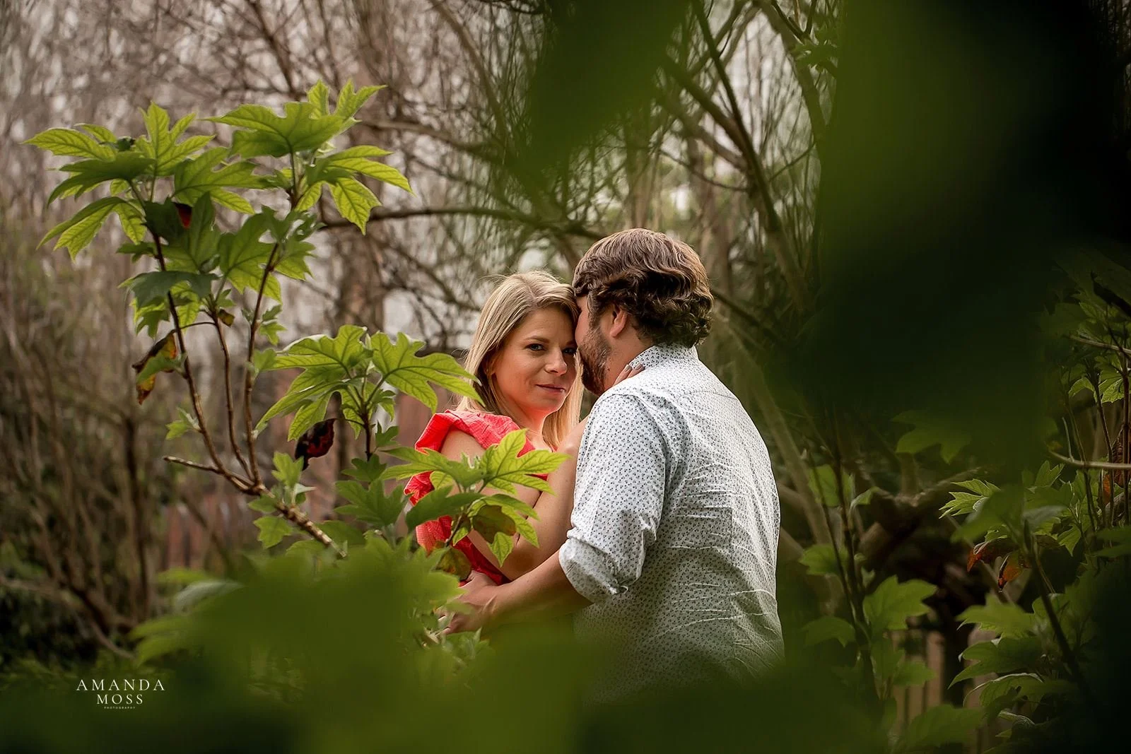 couple embraces while woman looks at camera through leaves in the foreground at rosies wine bar outdoor engagement session charlotte nc