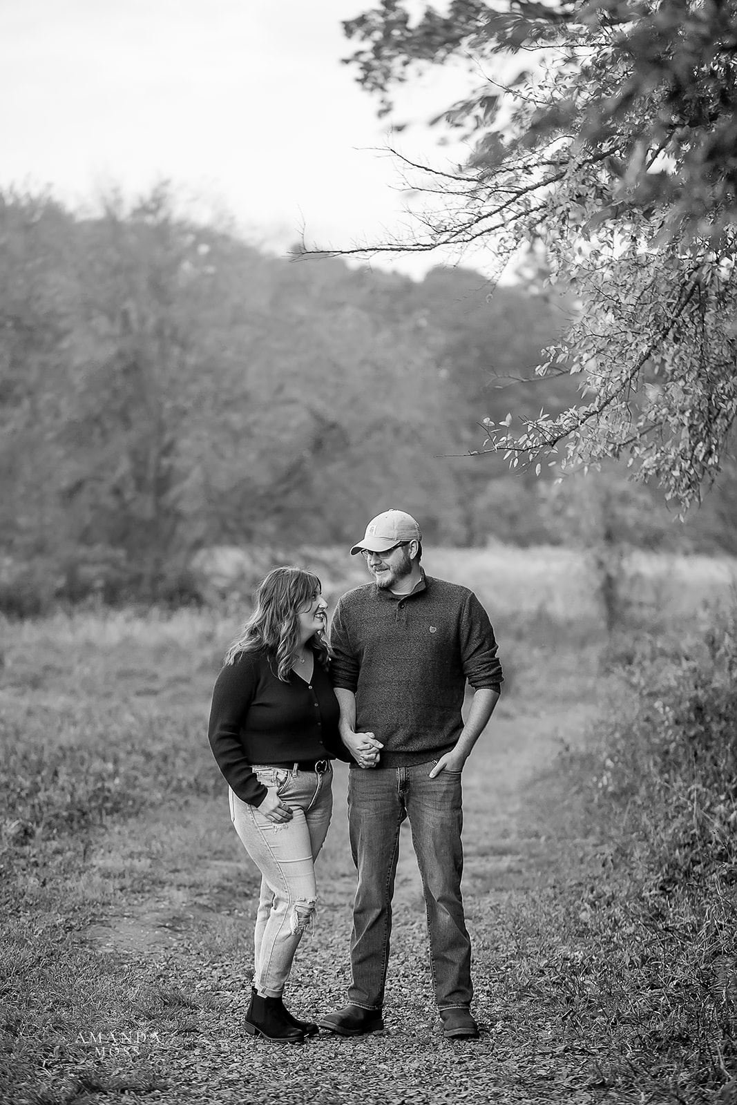 black and white photo of man and woman holding hands and looking at each other while standing on a woodsy trail