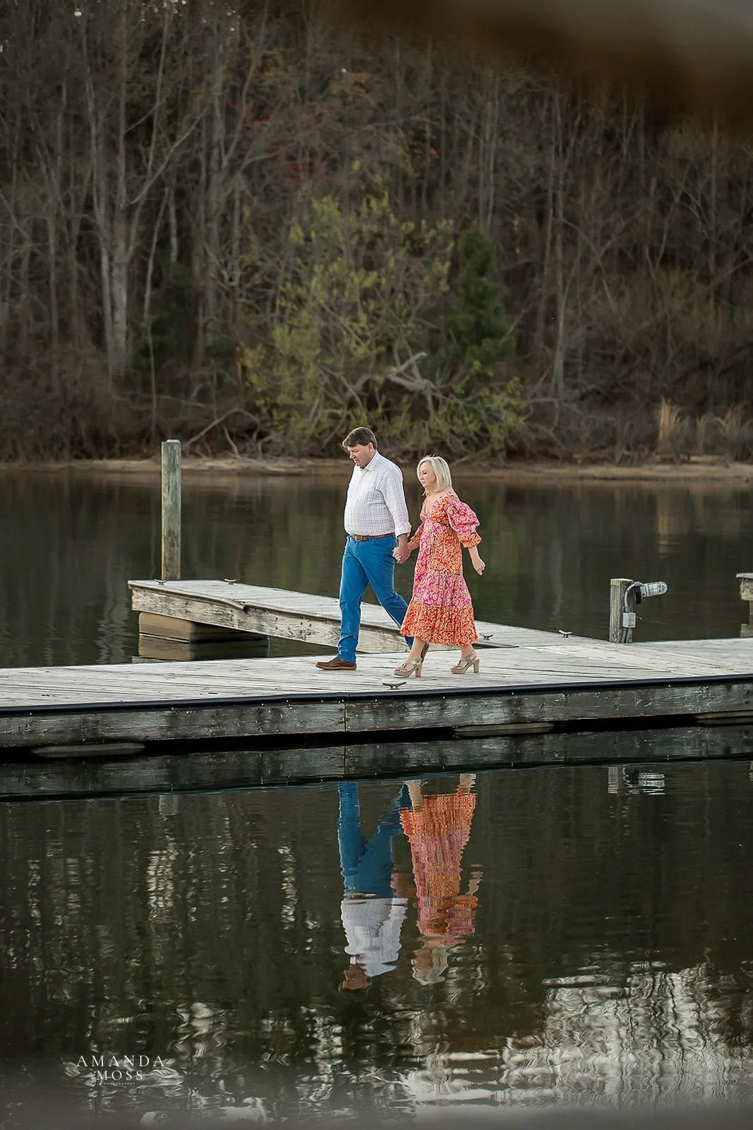 engaged couple walks across dock with their reflection in the lake at lake norman north carolina