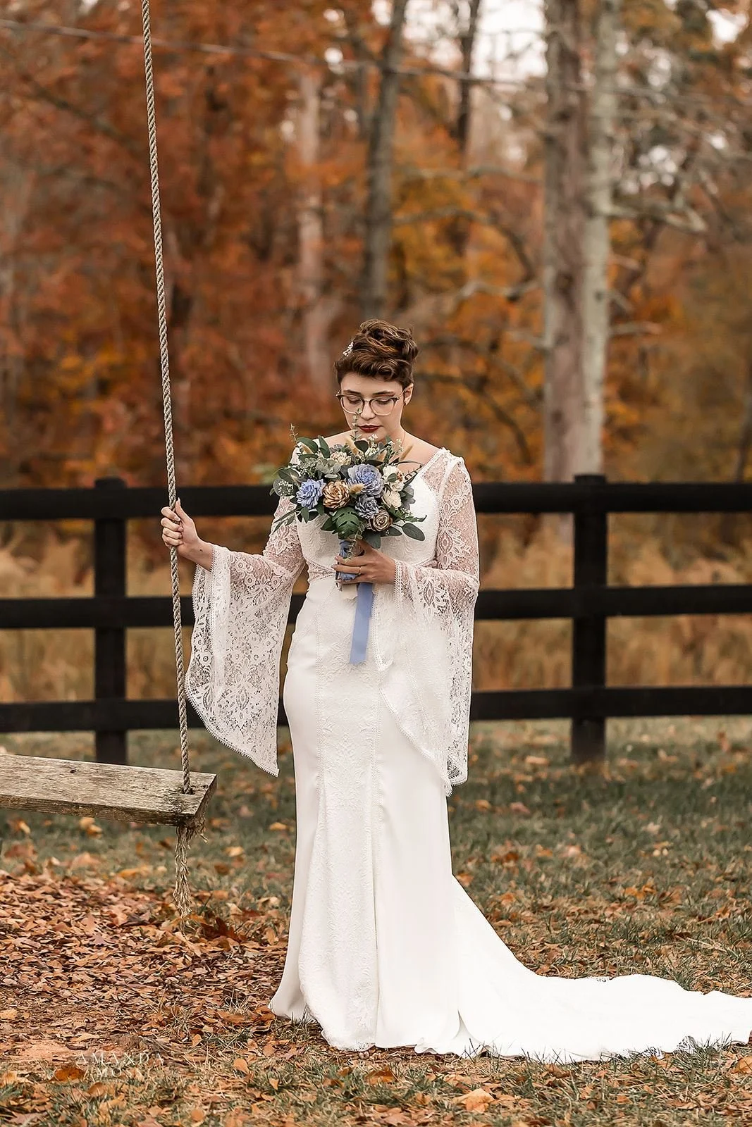 bride smells her bouquet beside a wooden swing in front of fall foliage at bansen farms wedding venue in north carolina