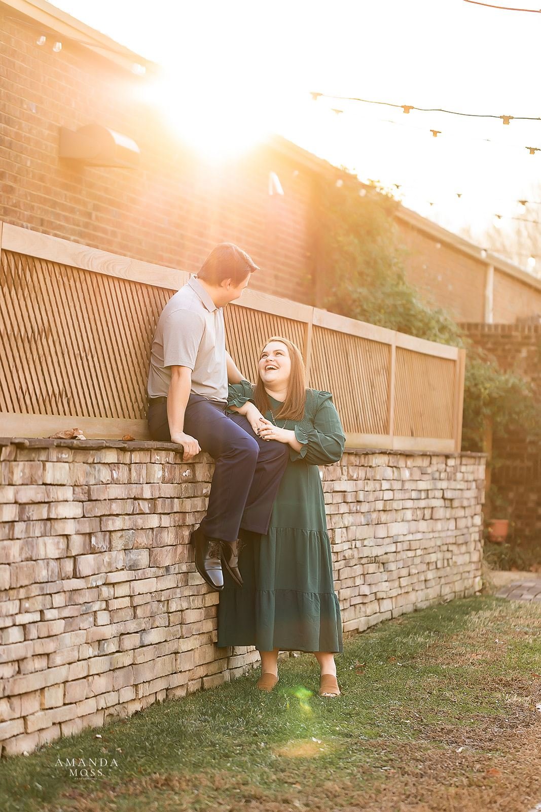 man sits on wall and laughs with his fiance in a green dress at magnolia woods in huntersville nc for outdoor engagement session