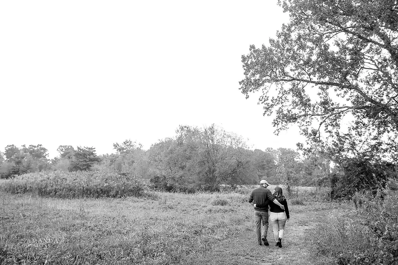 black and white image of a couple walking away from the camera into a woodsy field
