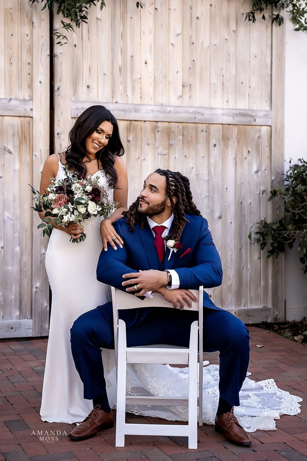 groom in navy suit sits on chair while bride holding bouquet stands next to him smiling