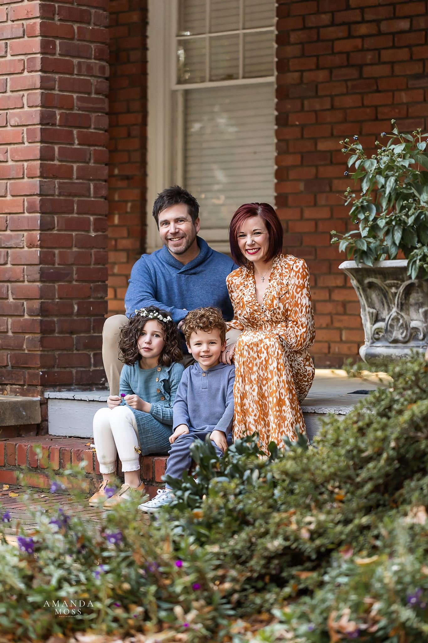 mom dad and two young children sit on porch of brick home in outdoor family photo session at glencairn gardens in rock hill sc
