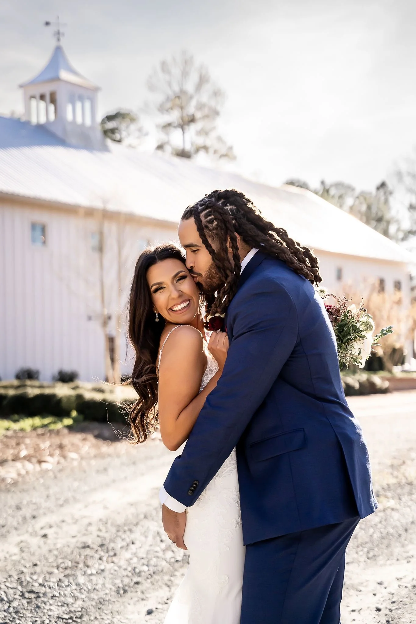 groom embraces his bride and kisses her on the cheek outside of the wedding venue barn of chapel hill