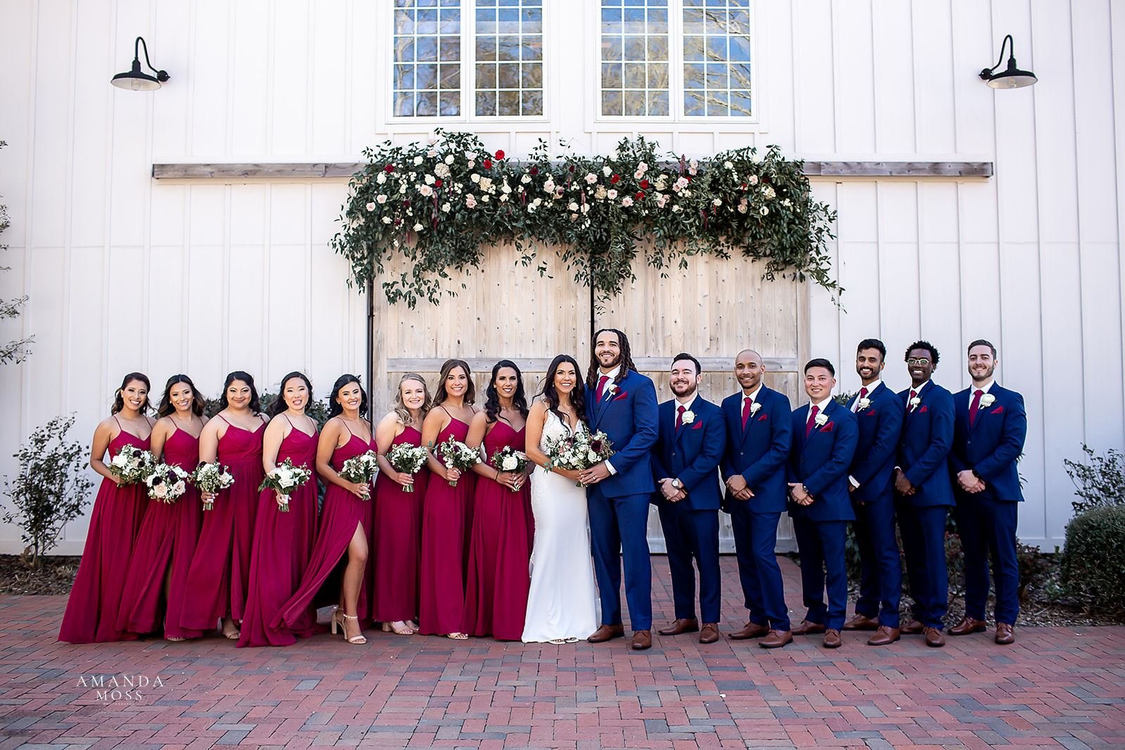 bride and groom with their wedding party in navy suits and burgundy dresses pose in front of barn of chapel hill