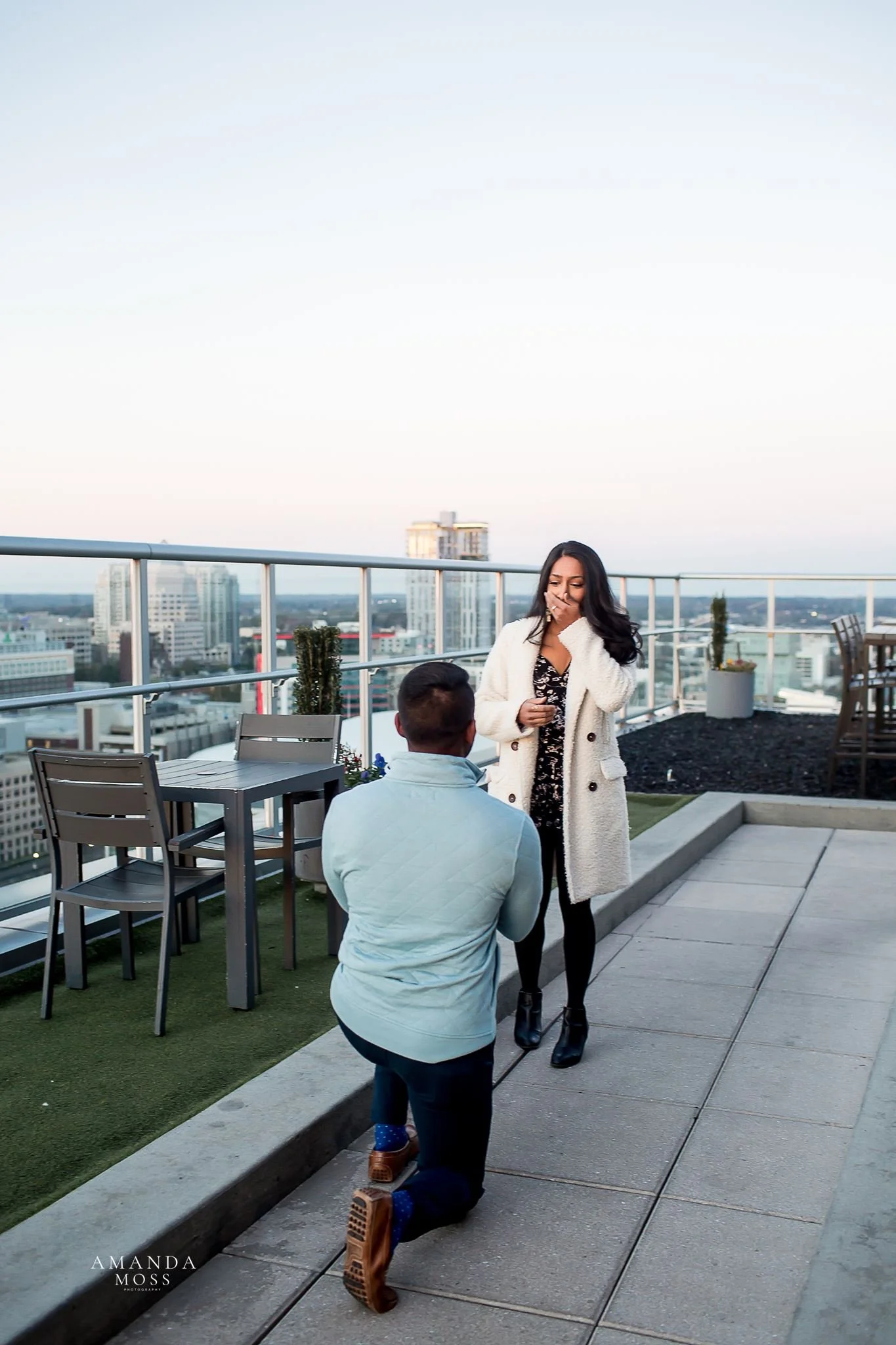 woman covers her mouth in shock while her boyfriend is on one knee proposing at fahrenheit rooftop patio