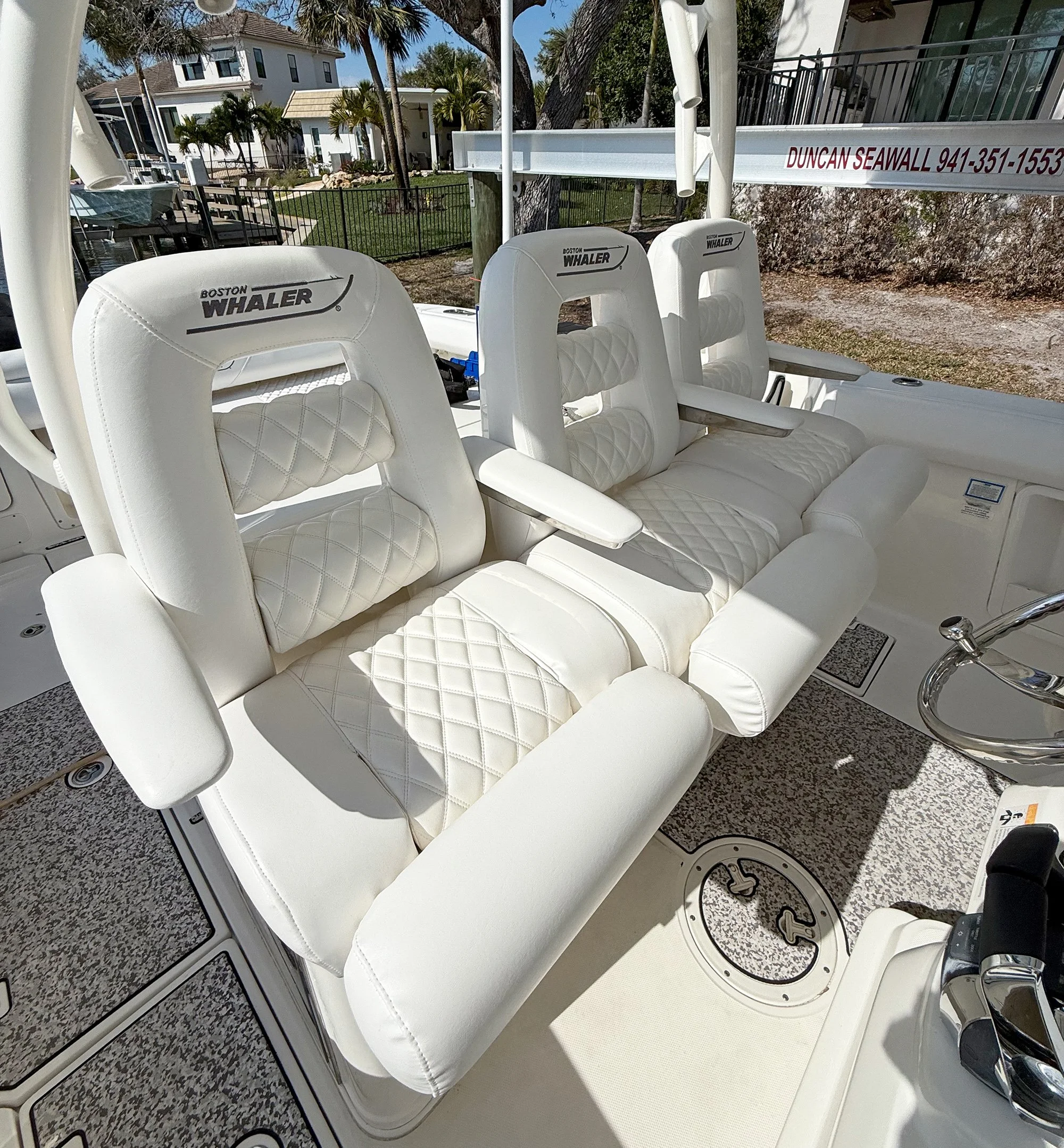 Close-up view of a boat's interior seating area with white cushioned seats labeled 'Boston Whaler' and a view of houses and palm trees outside.