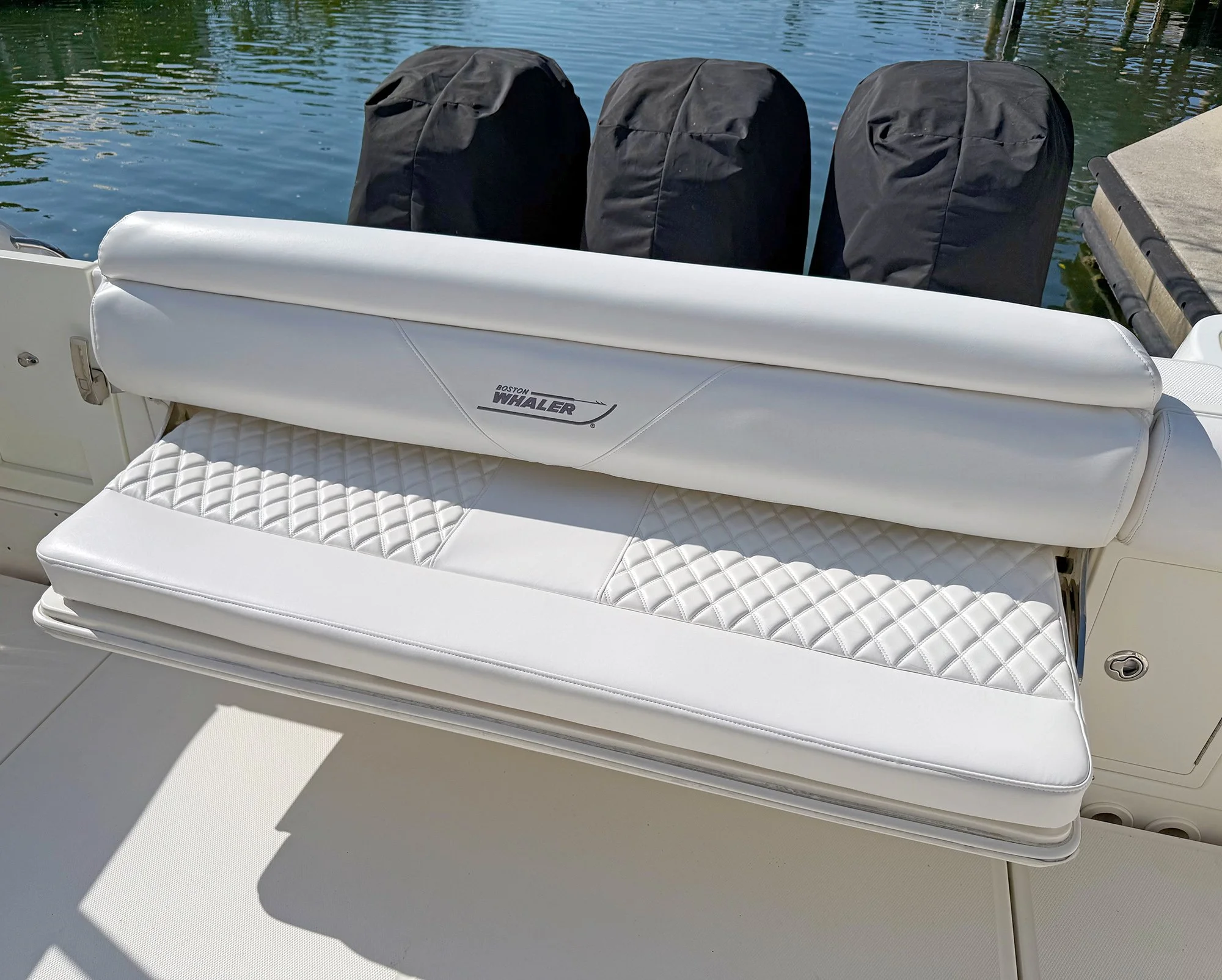 Close-up of a white boat seat and backrest with a stitched diamond pattern and a sticker that reads "Boston Whaler." Three black outboard motors covered with black protective covers are visible behind the boat, with a waterway in the background.