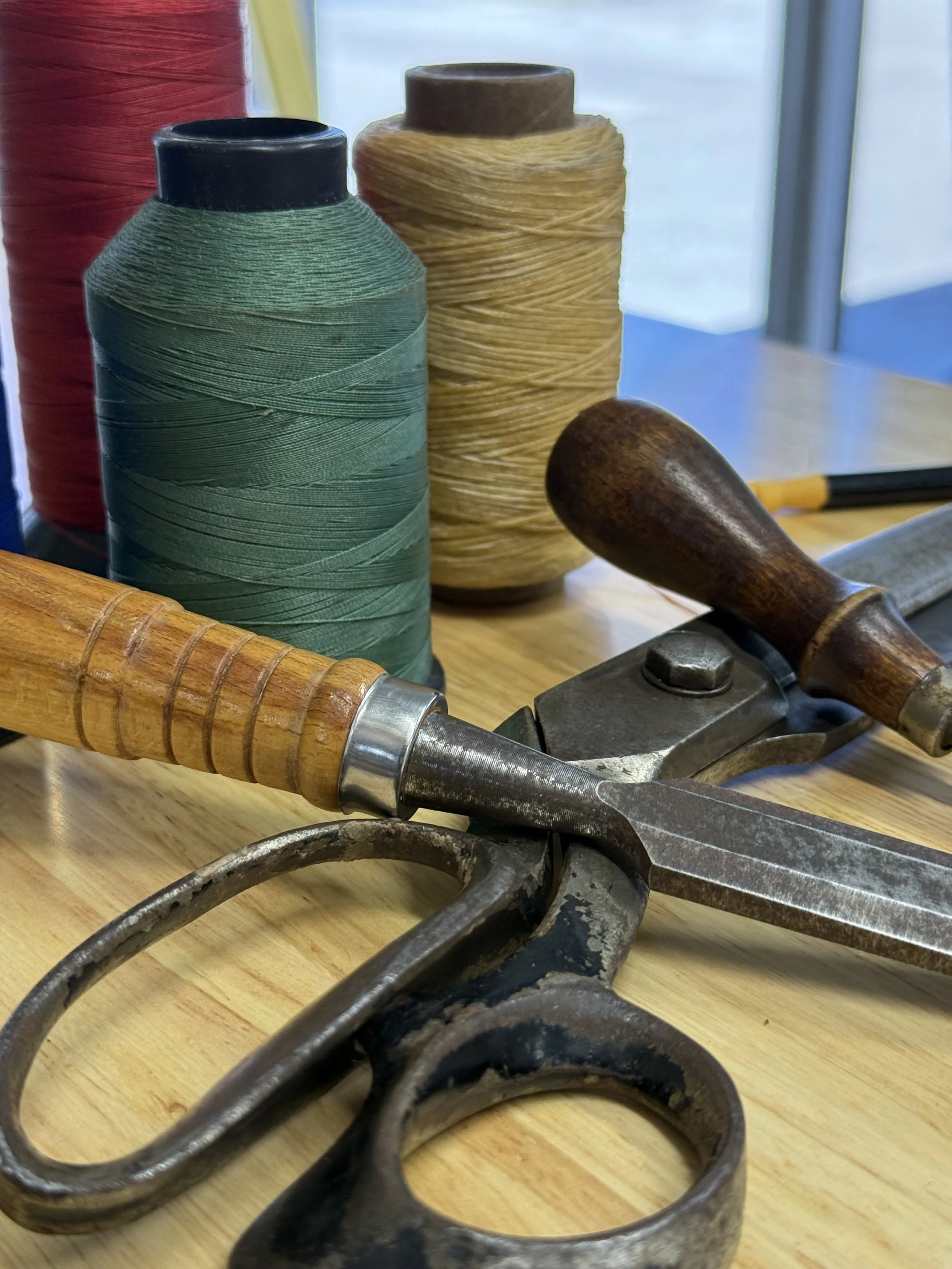 Spools of colorful thread or yarn in red, green, and yellow, along with vintage metal scissors and a wooden-handled tool on a wooden surface, with a glass window and outdoors in the background.