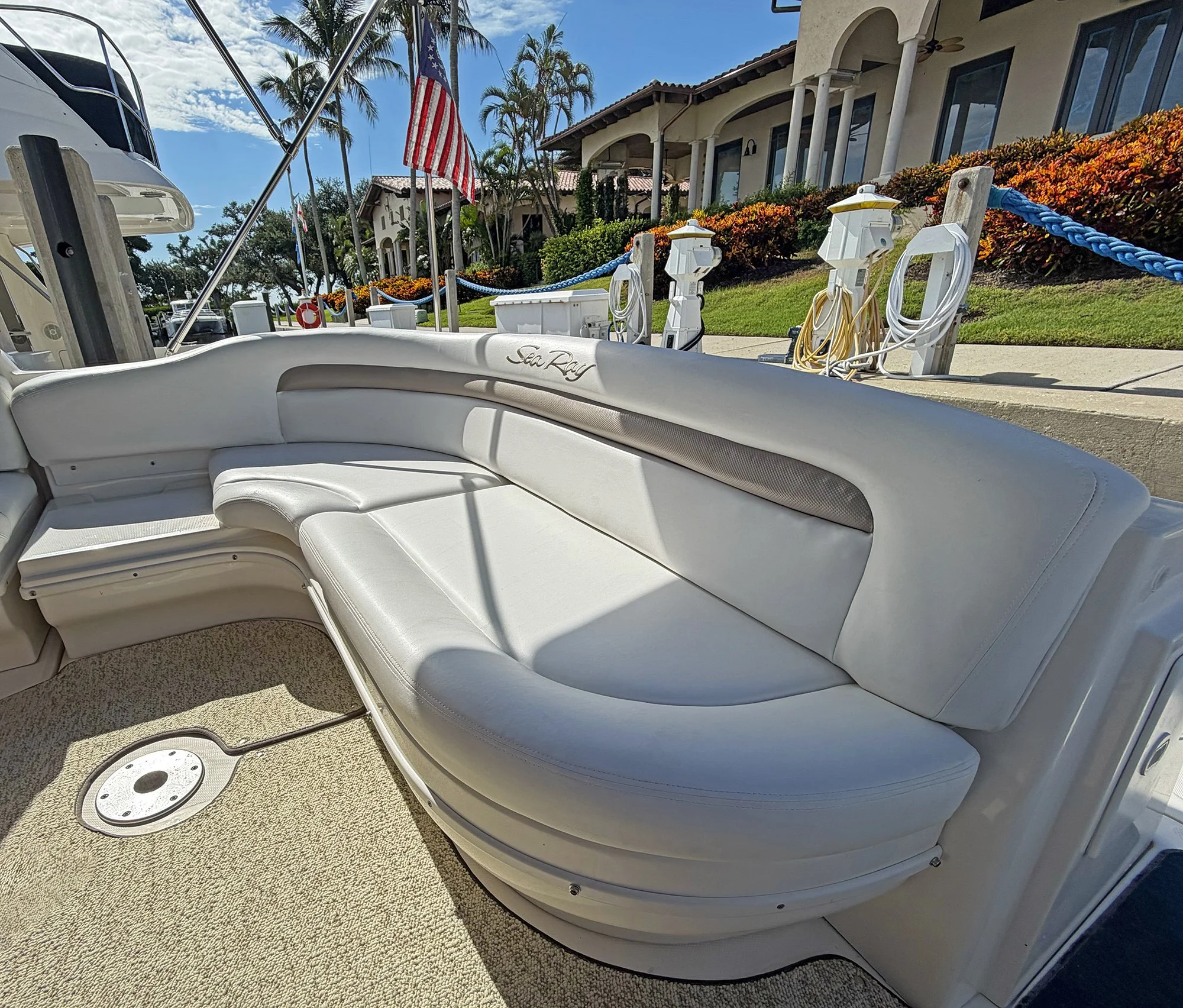 View of a white Sea Ray boat with electrical outlets and extension cords, docked near a house with tropical landscaping, palm trees, and an American flag on a sunny day.