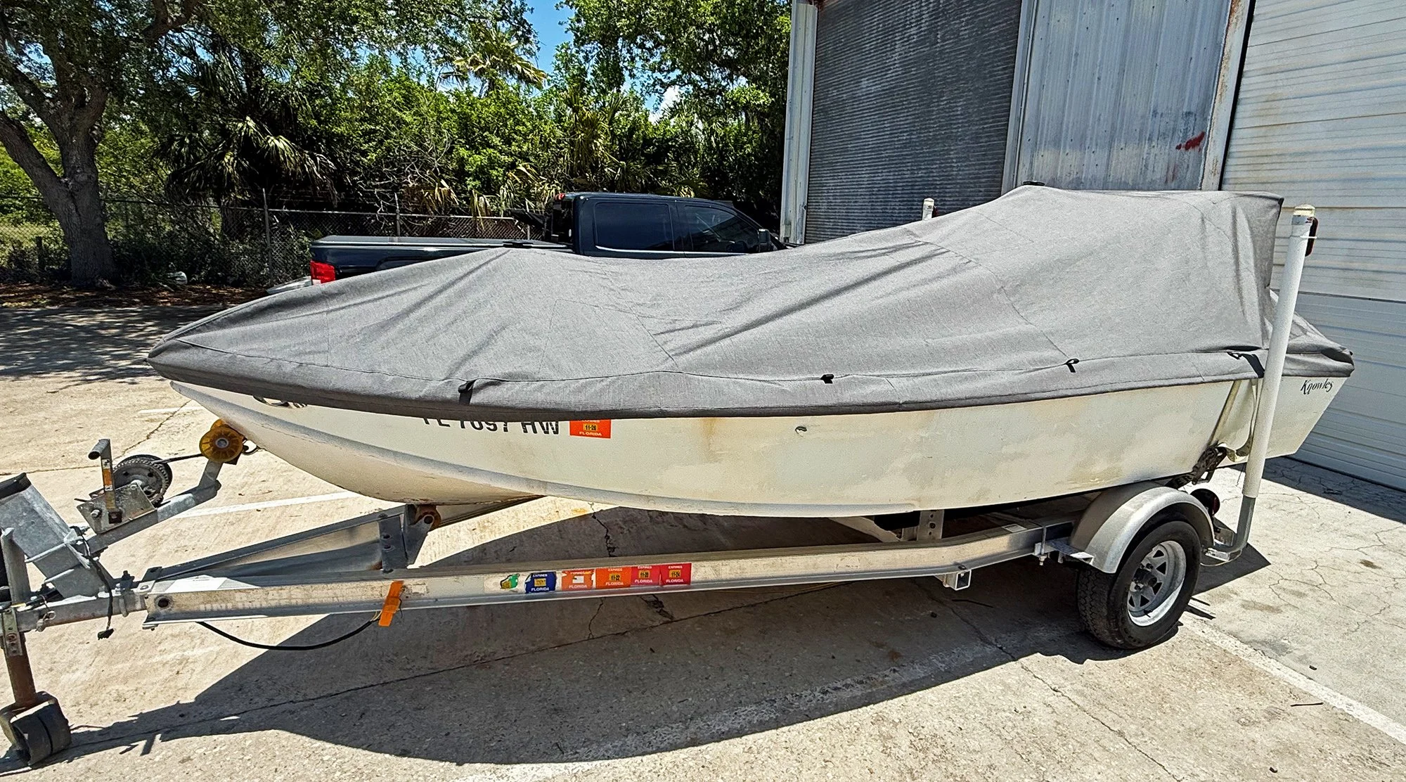 A small boat covered with a grey tarp on a trailer parked outside near a building and trees.