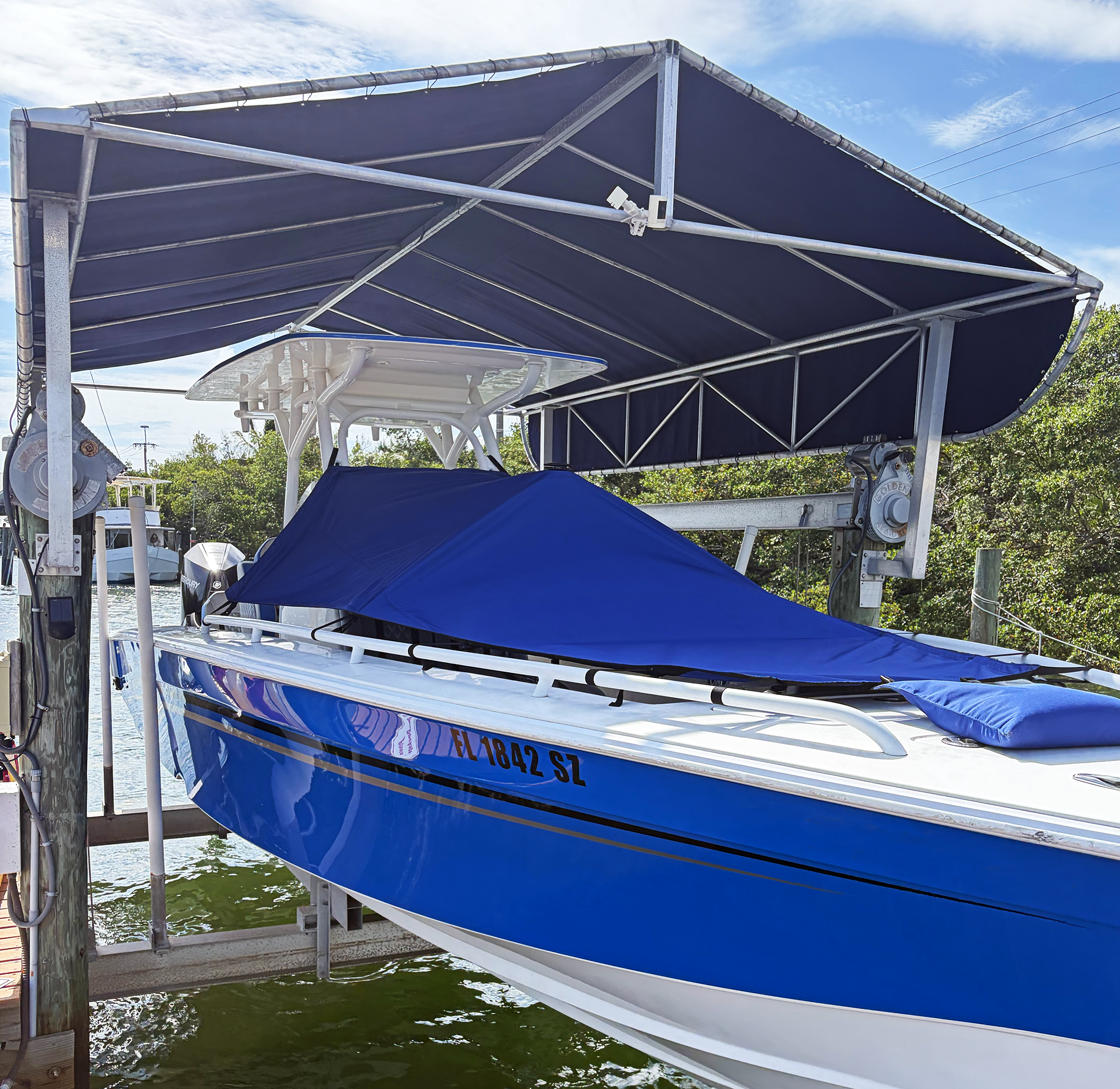 A boat lift with a blue boat suspended above water, with a metal canopy overhead and trees in the background.