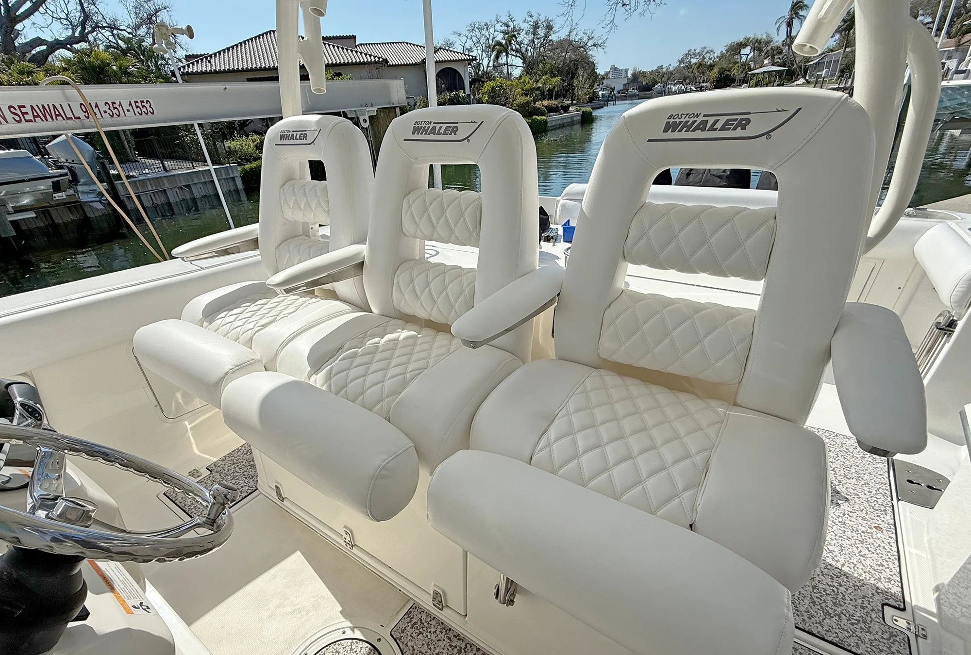 White cushioned boat seats with quilted patterns, labeled 'Boston Whaler', on a boat docked near a canal with houses and trees in the background.