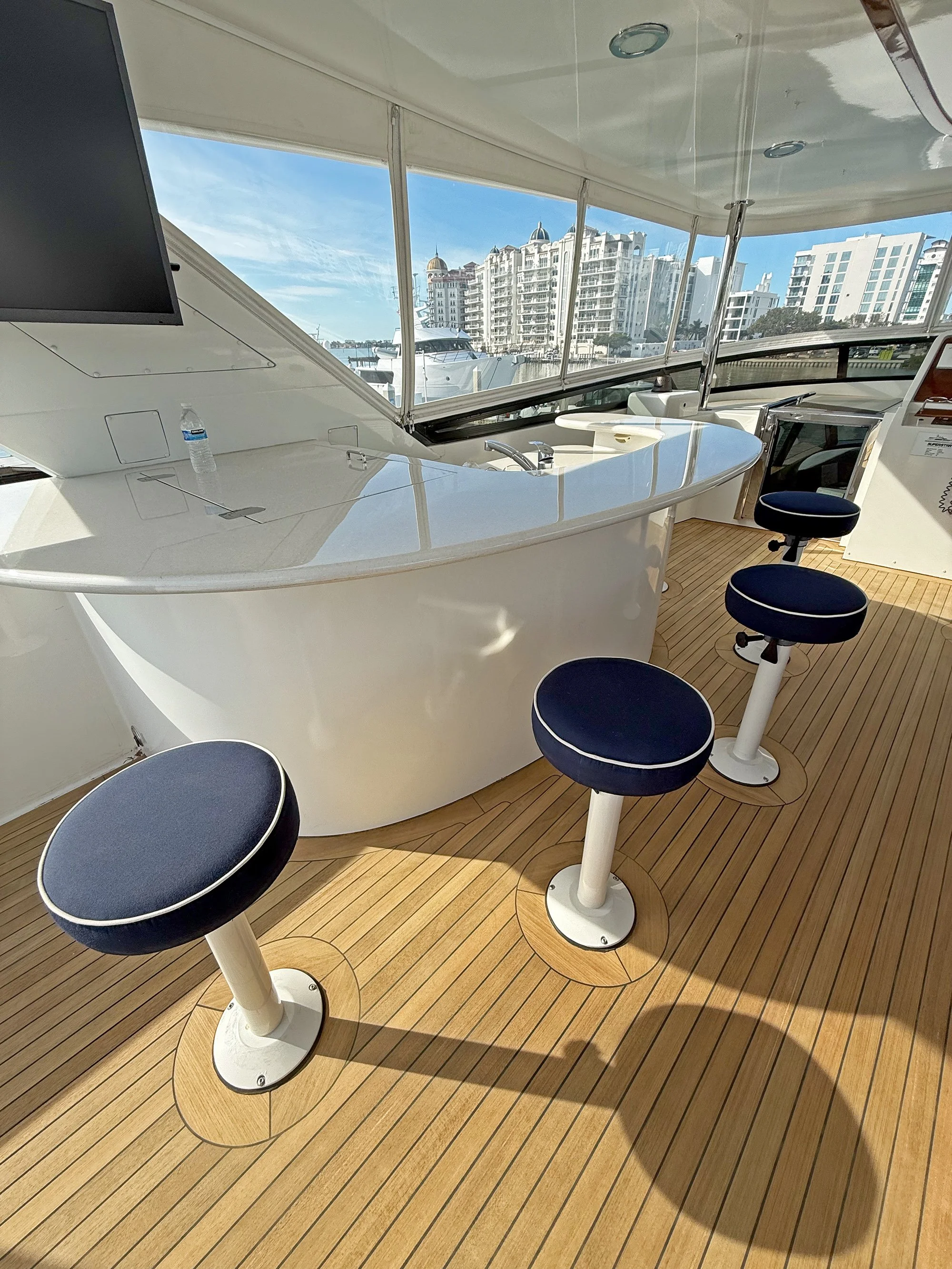 Interior of a yacht with a curved white bar, blue cushioned stools, a sink, a monitor, and large windows showing city buildings and boats outside.
