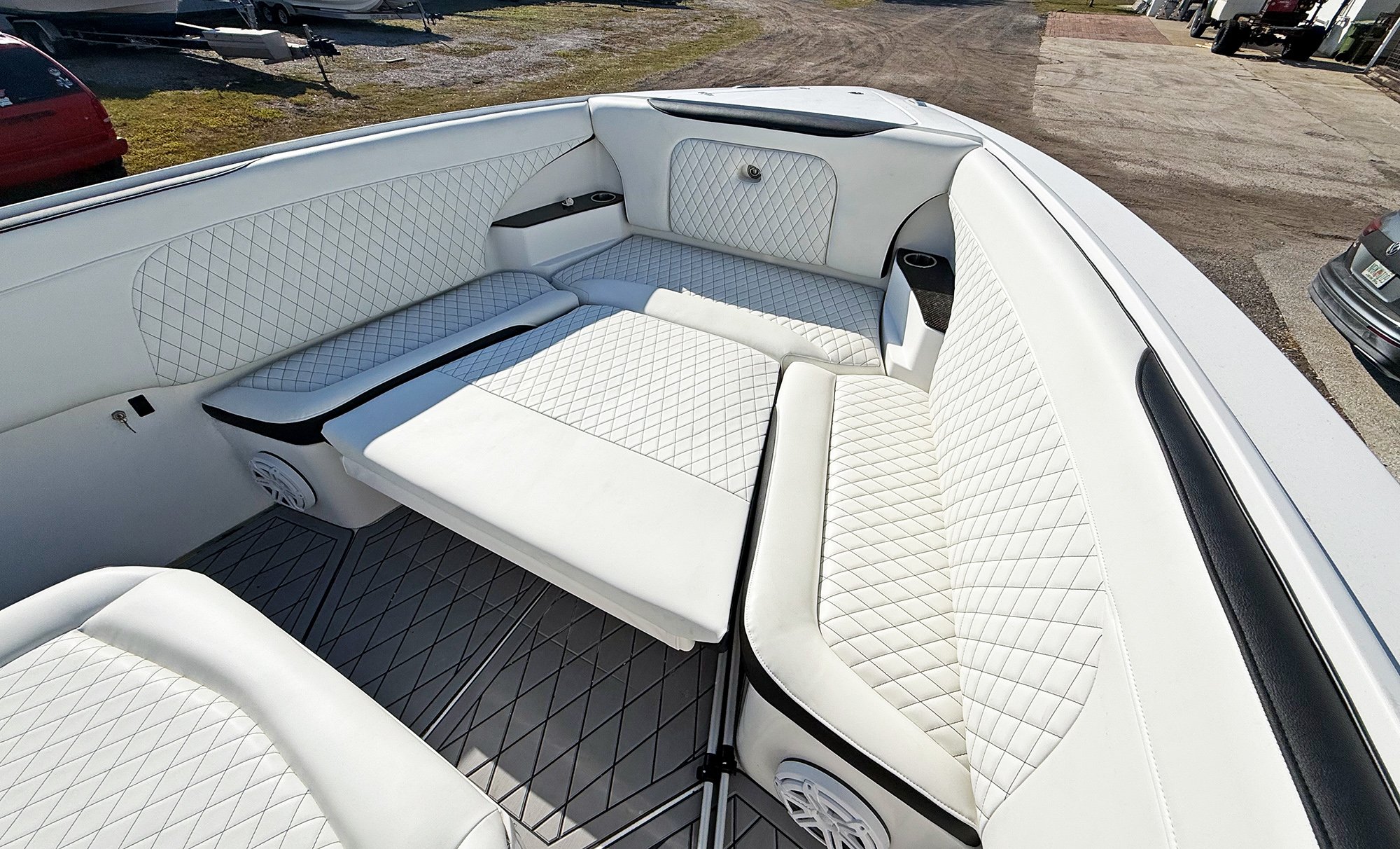 The interior of a white boat with white quilted upholstery, seating area, and built-in cup holders, parked outdoors on a gravel lot with other boats and vehicles visible in the background.