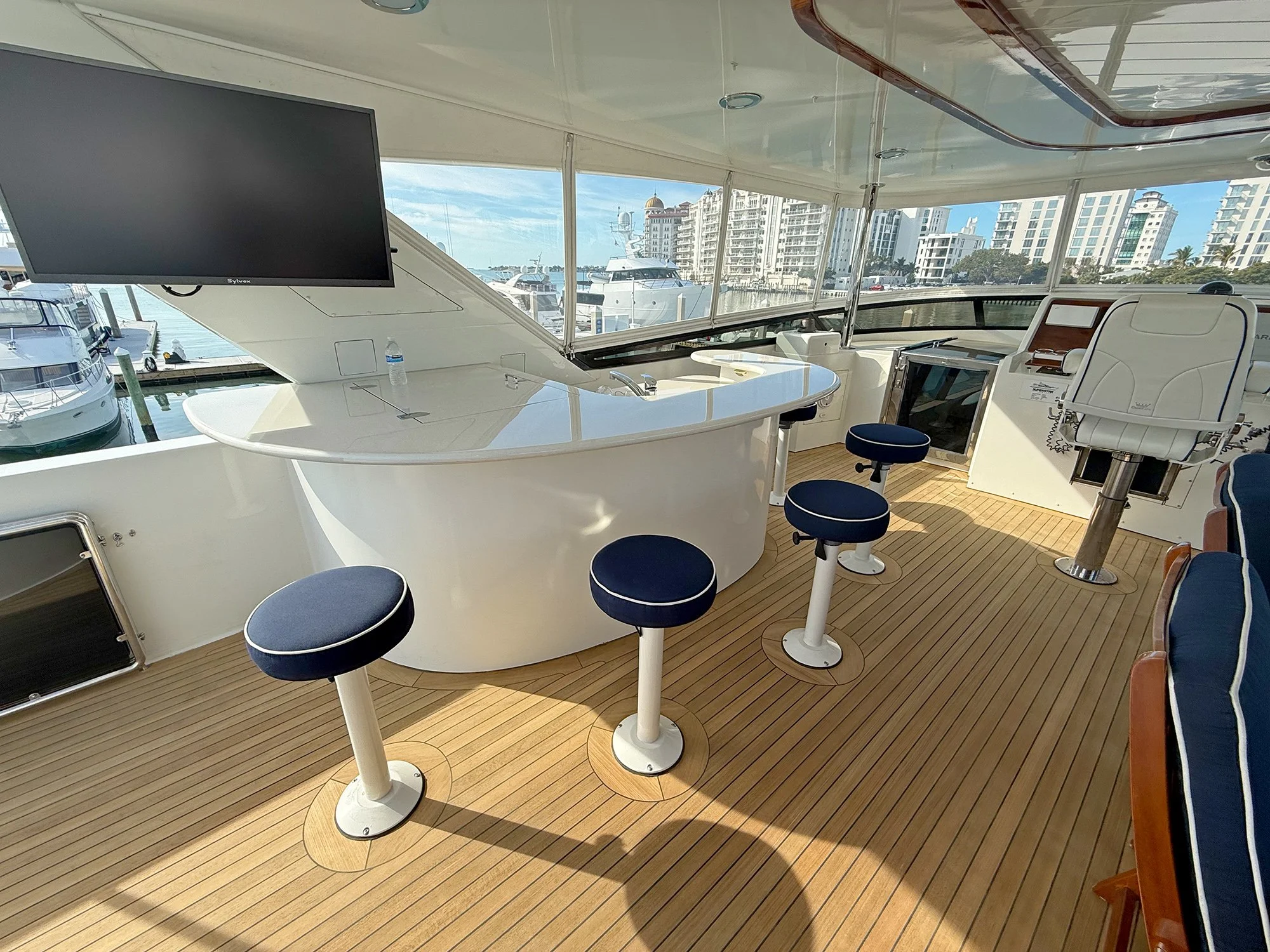 The interior of a yacht's enclosed deck overlooking a marina with boats and city buildings in the background. It features a white curved bar, multiple blue stool seats, a mounted flat-screen TV, and wooden flooring.