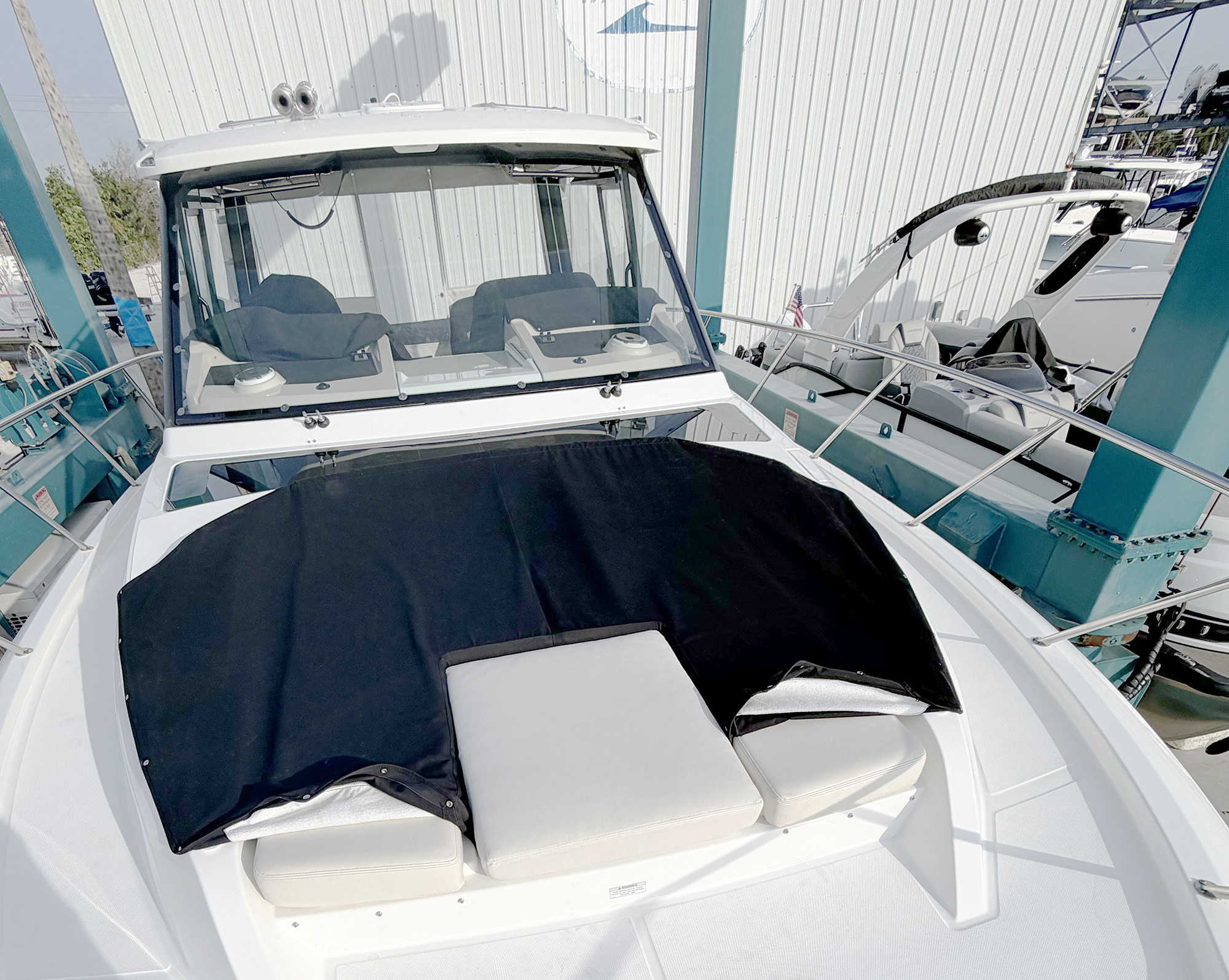 A white boat with cushioned seating and a black cover, docked at a marina with other boats and a large metal building in the background.