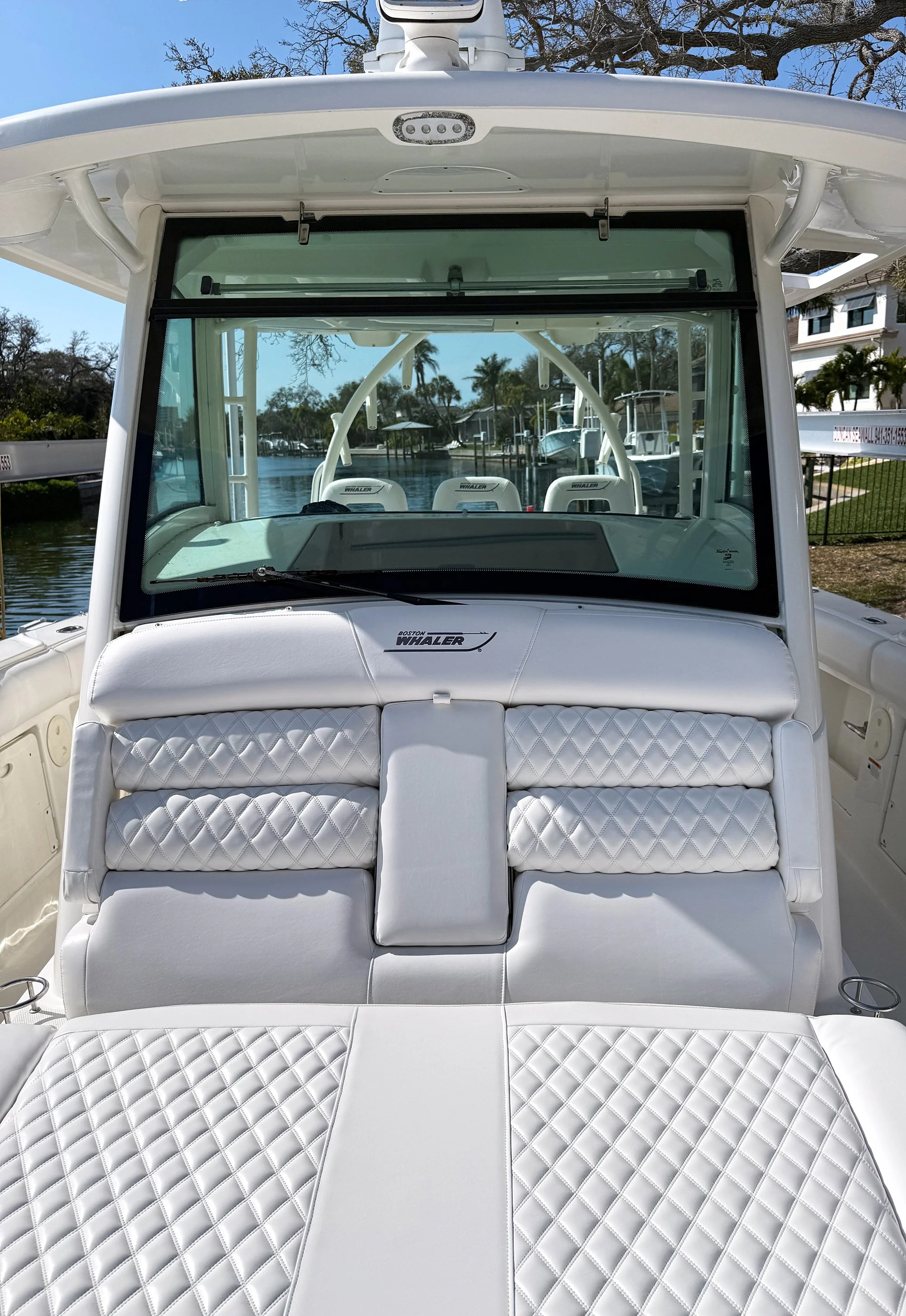 The image shows the front view of a white Boston Whaler boat with cushioned seating. The boat is docked on a calm waterway with houses and palm trees in the background.