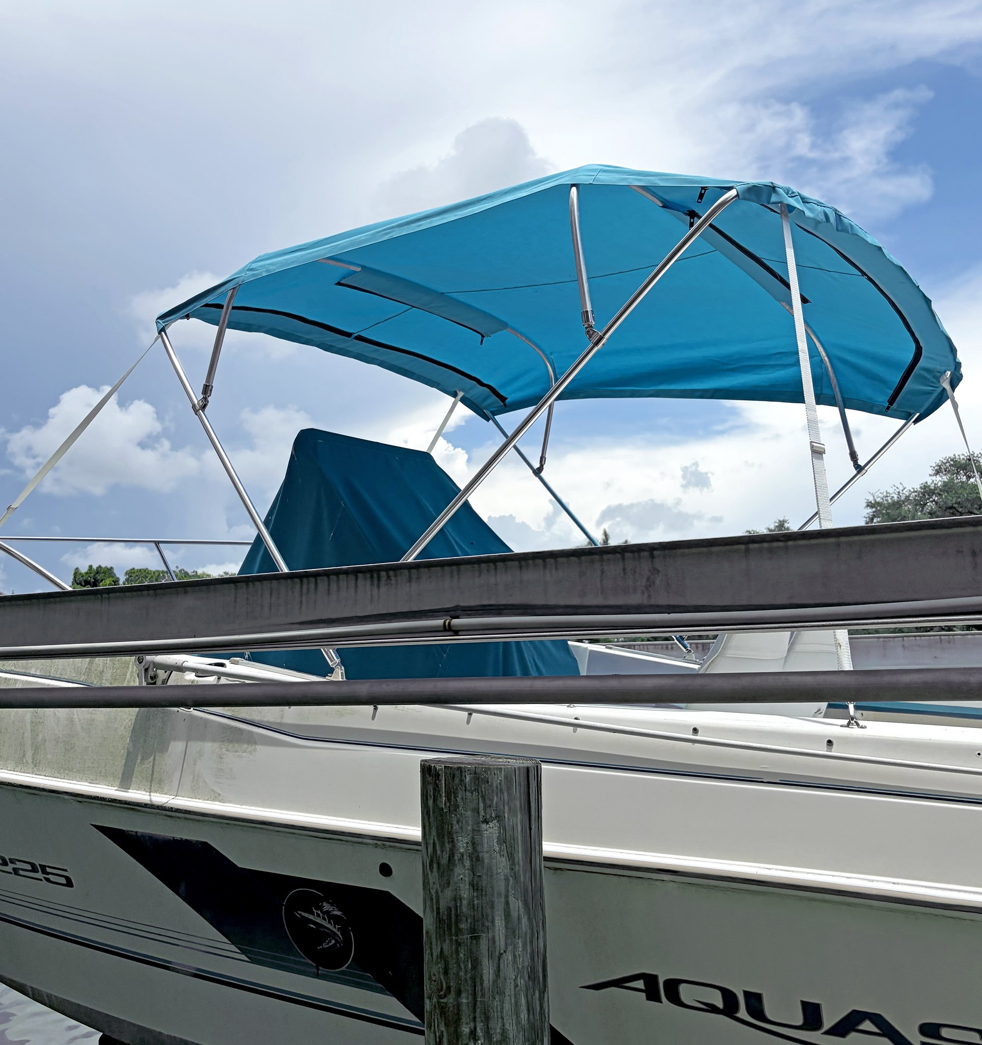 Photo of a blue boat canopy supported by metal poles, with part of a boat visible below and a wooden post attached to the boat, set against a cloudy sky.