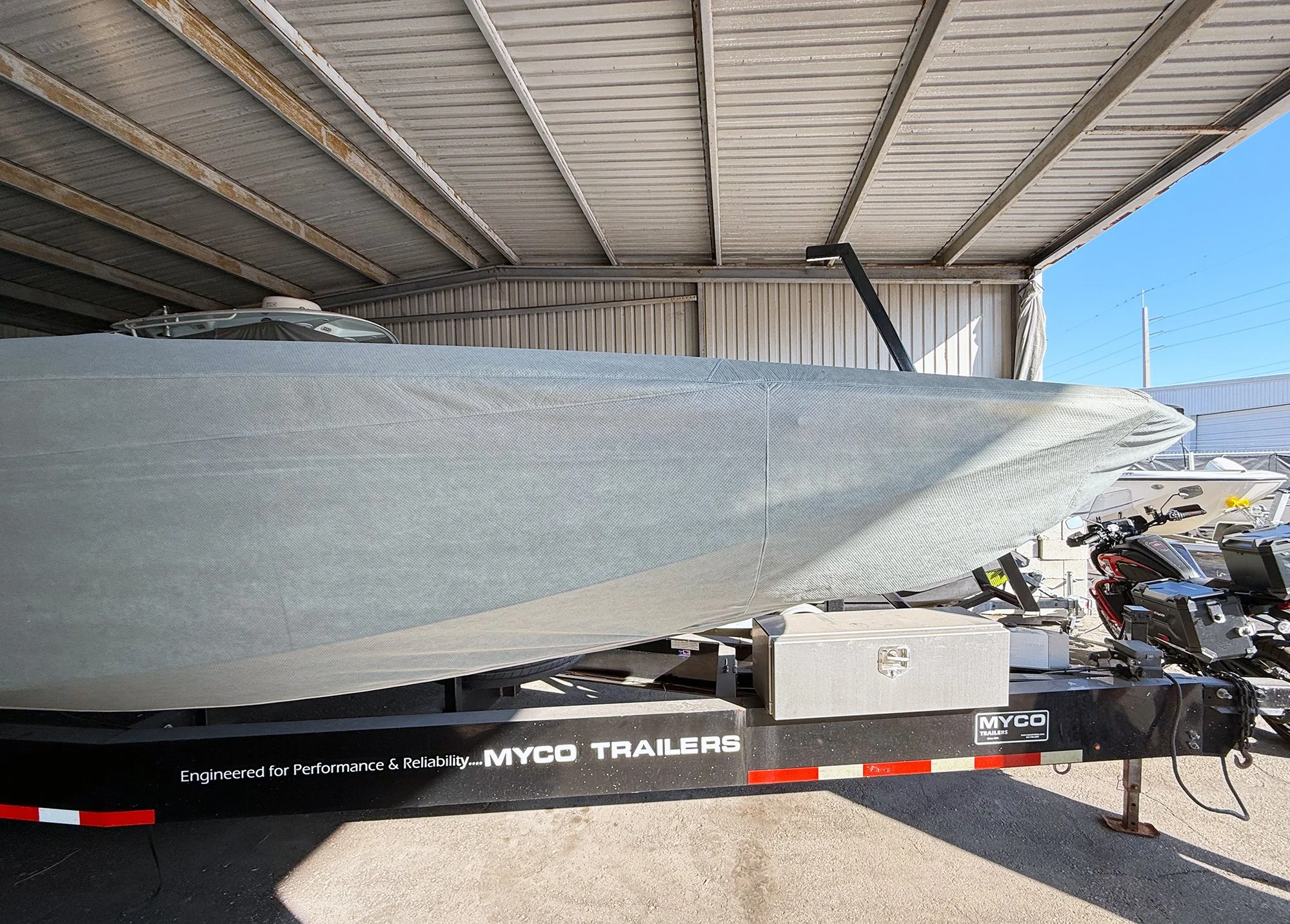 A boat on a trailer parked under a metal roof with blue sky visible in the background.