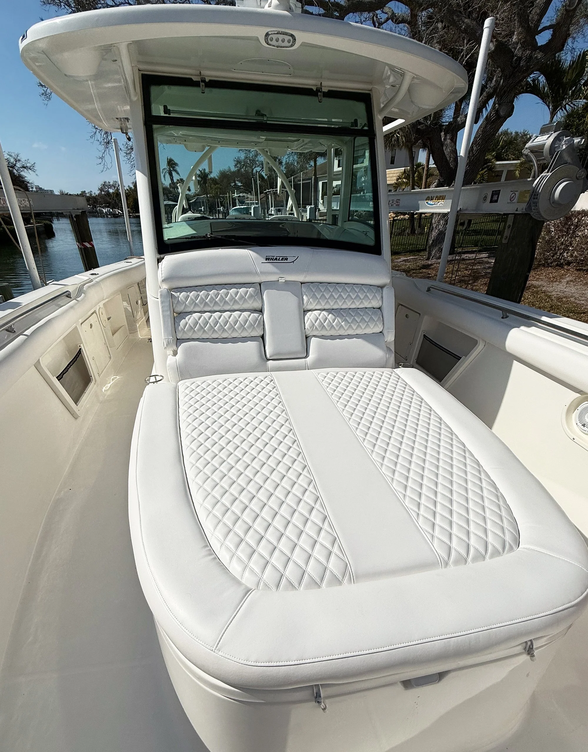White boat with cushioned seating area and a glass windshield, docked by a marina with trees and a fence in the background.
