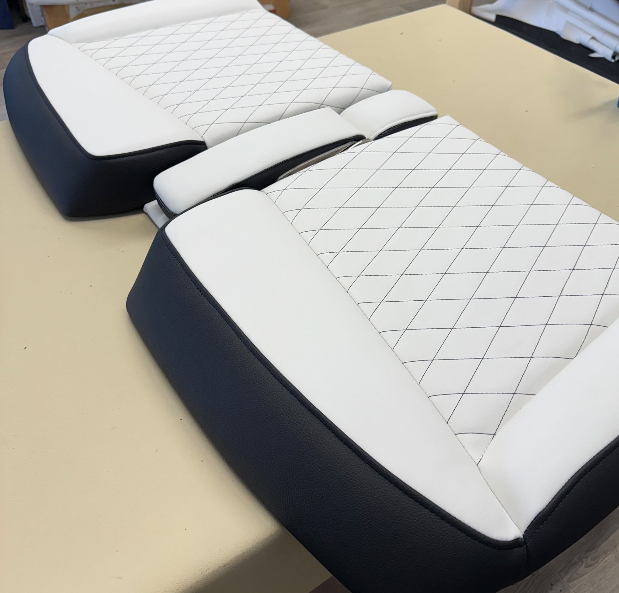The image shows a white and black car seat with diamond-patterned stitching, placed on a beige surface. In the background, there are shelves with various objects and a workspace setting.