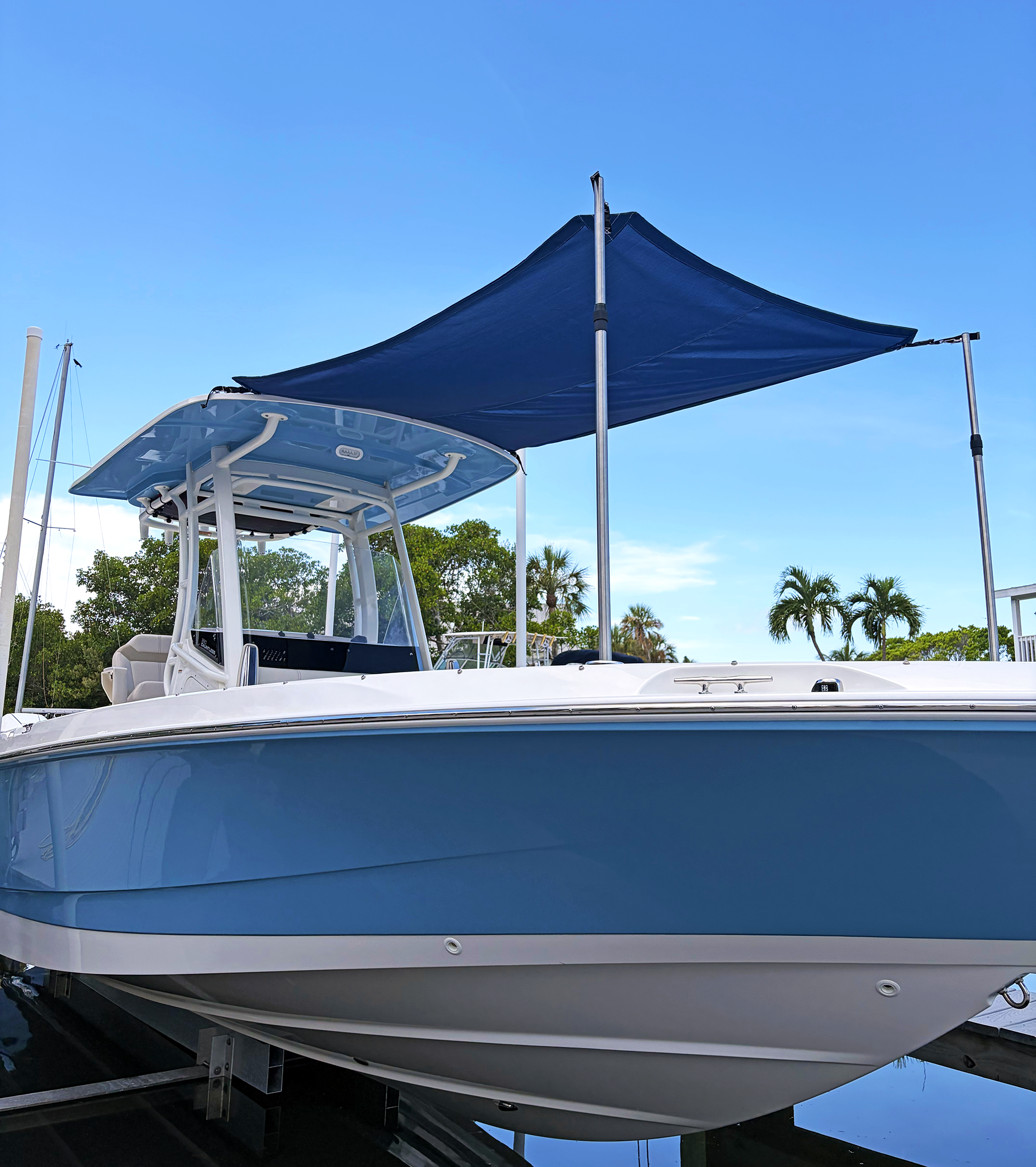 A white yacht boat with a blue canvas shade structure on top, docked at a marina with a clear blue sky and palm trees in the background.
