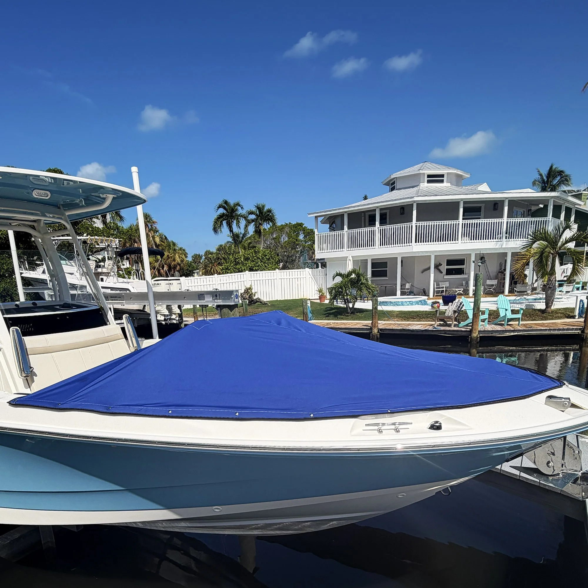 A white boat with a blue cover docked in a marina, with a white house, palm trees, and a blue sky with a few clouds in the background.