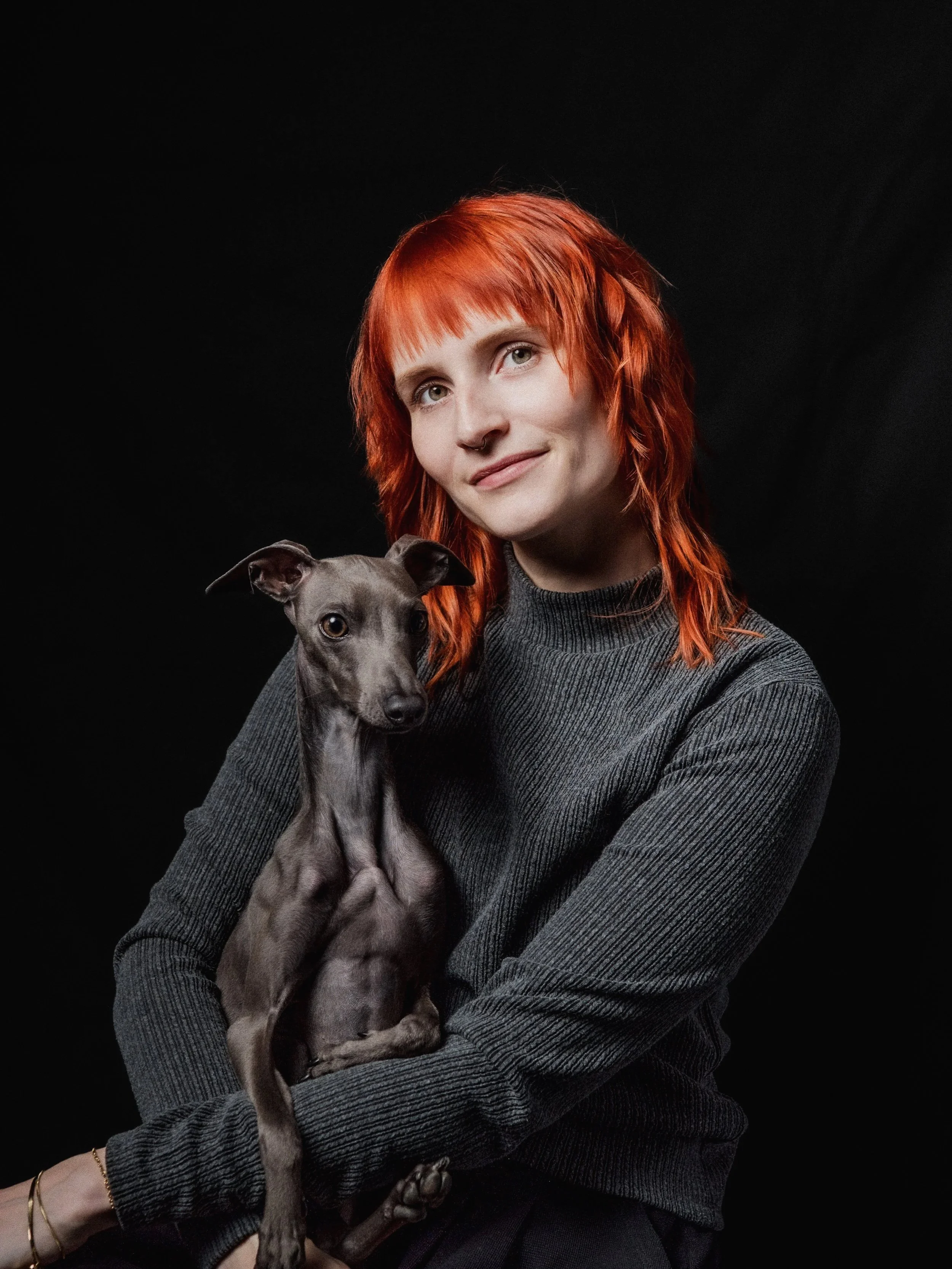 Eine Frau mit roten, zerzausten Haaren hält einen grauen Hund mit langen Beinen vor einem schwarzen Hintergrund.