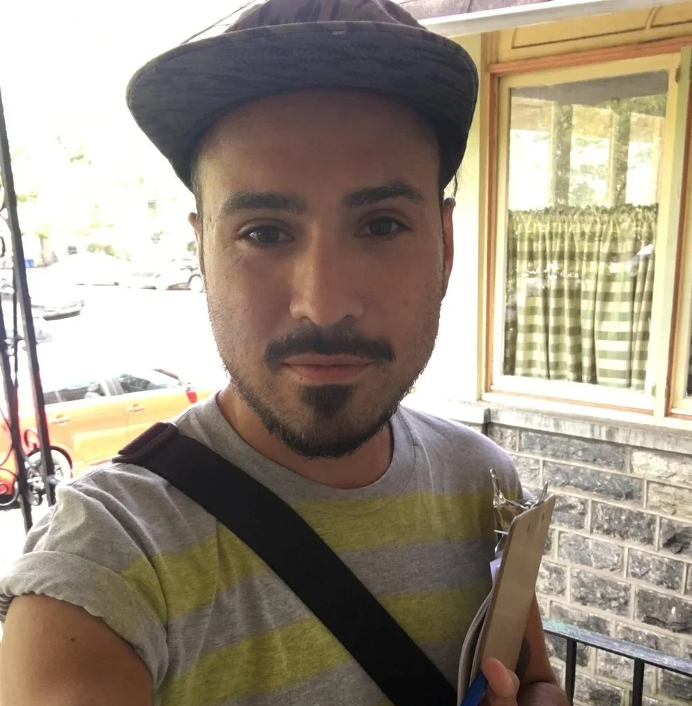 A young man with a beard and mustache wearing a striped t-shirt and cap, holding a clipboard and standing outside near a window with a yellow and green checkered curtain.