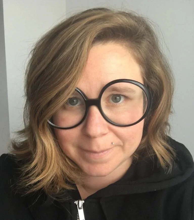 A woman with wavy light brown hair wearing large black round glasses and a black hoodie, smiling slightly at the camera.
